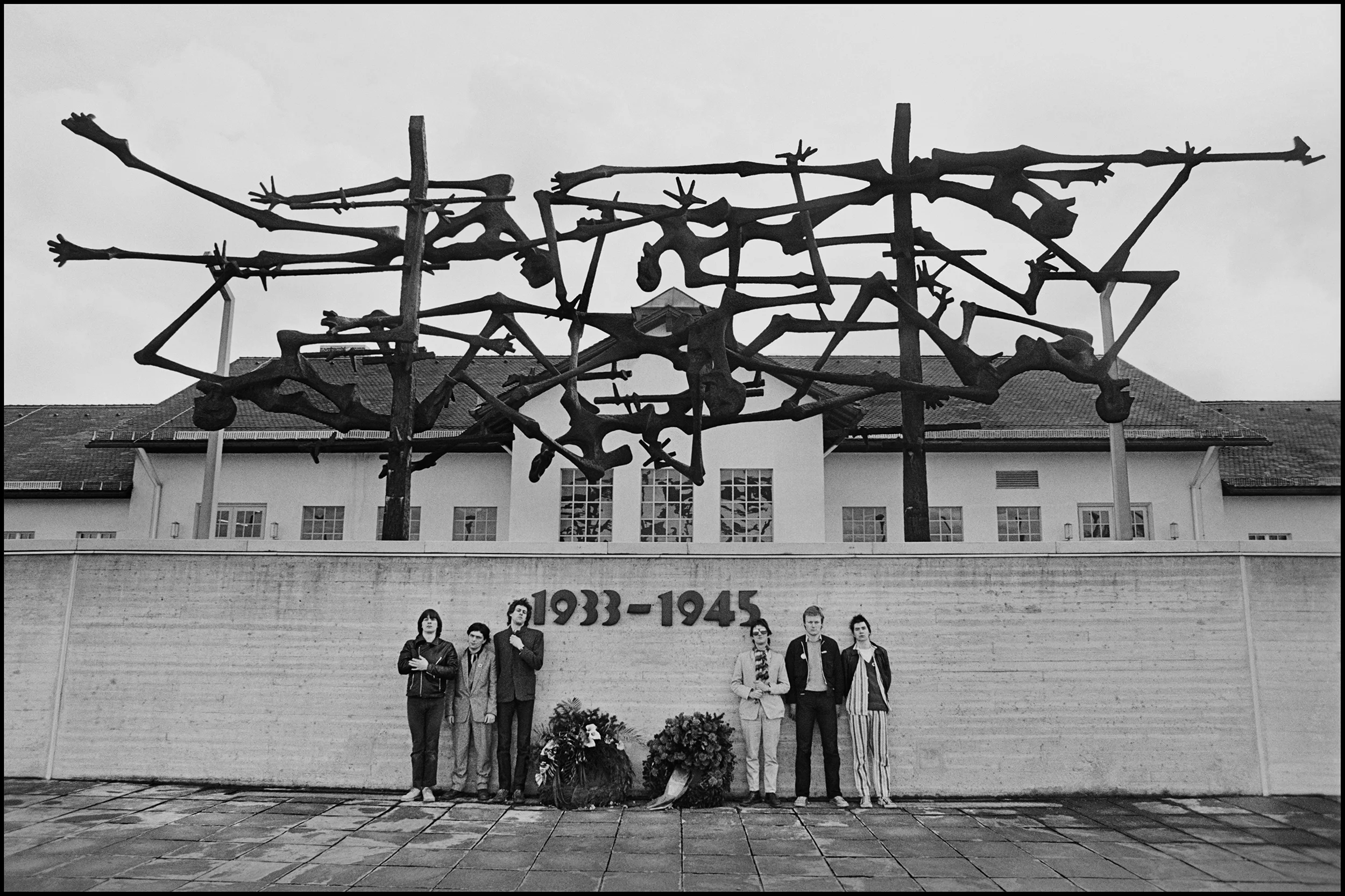 Boomtown Rats - Dachau Concentration Camp 1978 Pretty much every Tuesday you could board the 9am British Airways flight to Munich and find it full of Bands going to do the German TV Show Rockpalast which was broadcast live each week. I went along for the ride as there was often a spare ticket to be had. I had known Bob Geldof and the Boomtown Rats since their first single, they opened for Thin Lizzy at Dalymount Park and Geldof was a frequent visitor to the house I shared with Phil Lynott. By this time they were very successful and would do pretty much anything for a Photo, we went out to the Olympic Park to shoot stuff, it's where the Israeli Athletes were shot and although interesting as a background it put us in a somber mood, when we were done I figured we should go out to Dachau and visit the Concentration Camp, we had plenty of time and I thought it was something we should all see. I had no intention of taking photographs, especially as Johnny Fingers only wore pajamas and its hardly the place for a shoot, but, when I saw the giant sculpture over the gates I felt so overwhelmed by it that I asked the Rats to stand under it to give it some scale, just look at the emancipated people portrayed in it and the sheer power of the work. Makes Rock and Roll seem so inconsequential really, don't you think...
