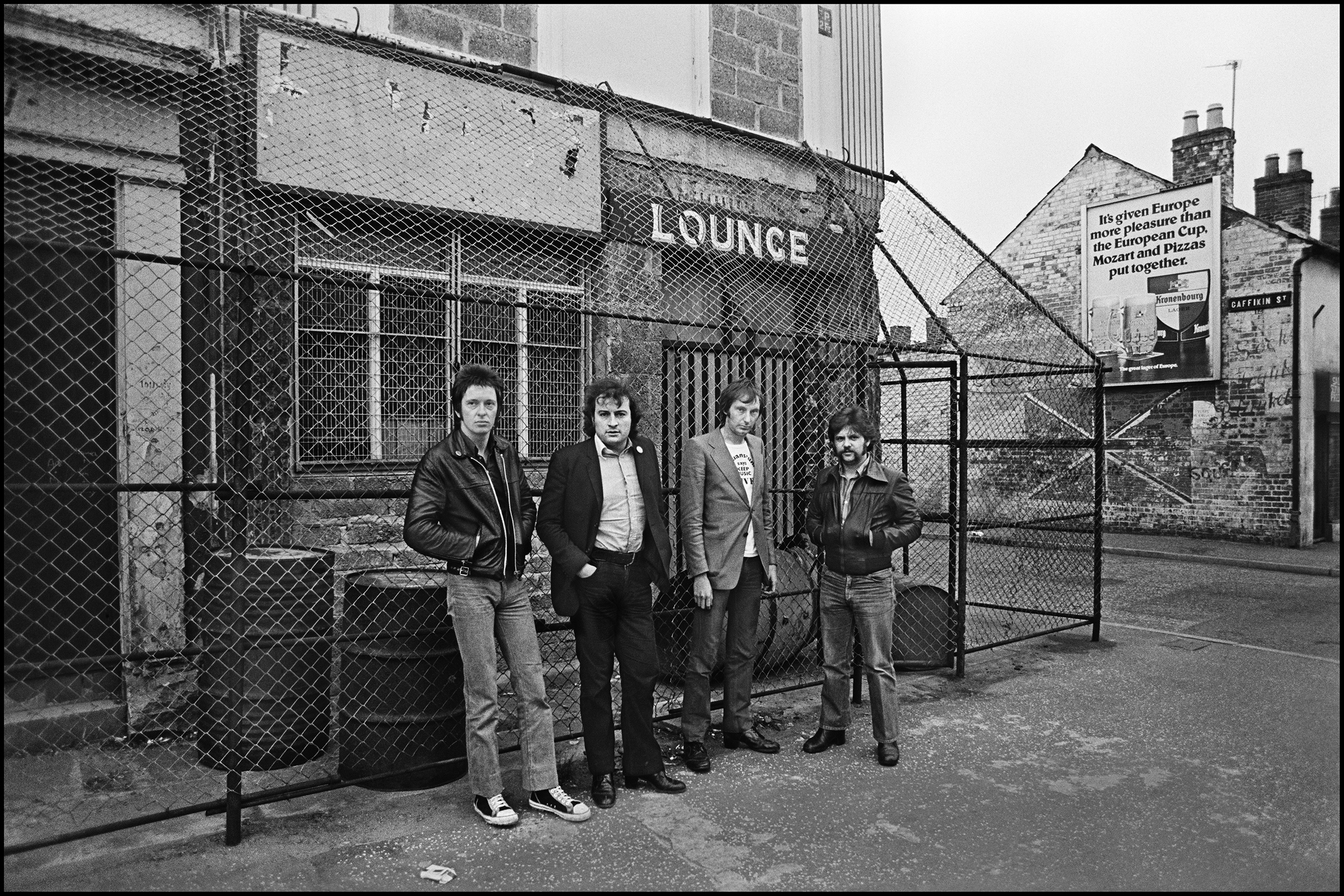 Dr Feelgood in Belfast 1978 The Feelgood's were pretty much the NME house band back in the mid seventies, we would review them at any opportunity and I had become friends with them via Jake Riviera. They had an animal intensity about them, but also a purity, a raw, sexual, hard and intense kind of purity. Lee in his white suit doing the splits and jerking off with the microphone, Wilko machine gunning the audience with his guitar and zig zagging around the stage like a maniac, plus with Sparko on Bass and the Big Figure on drums they had a magnificent and stoic rhythm section. A couple of the most memorable gigs were actually parties, Led Zeppelin made the mistake of inviting them to play the post gig party they threw after the dates at Earls Court in 1975. Playing in quite a large room somewhere in the depth of that huge semi deco building that bands like the Zep's and Floyd somehow made intimate, the Feelgood's turned a lot of heads away from the bar, it was a short set but a lively one, I often wonder if the short set was because Peter Grant, Led Zeppelin's manager did not want people to forget the real reason people were there, to see Page and Co give an American sized show in the UK. Plant preening and Page with his Violin bow and laser was impressive up to a point, and eighteen thousand punters went home happy but it was the other four piece, the ones from Canvey Island I enjoyed the most, they played their hearts out, just like they always did. They also played the NME party in 1976 at Dingwalls, packed to the rafters they ripped the place up with their own brand of Rock and Roll. That night Ian Dury arrived with a razor blade on his earring, Punk was just around the corner. They also played the benefit to Save the Hope and Anchor, an awesome venue in North London, playing in a basement with sweat dripping off the ceiling, it was so crowded you could hardly breathe, they put in one of the best live shows I had ever seen. Thanks to Jake I often went to Feelgood's gigs when there was a spare seat in the bus or on the plane, one time we flew to France and came straight home in this little light aircraft, not even bothering to spend the night there. Despite knowing them so well I only shot them offstage the once, and sadly this was after Wilco had left the band. It was in Belfast, and I was not really prepared for the shock of seeing the true side of Northern Ireland. Nothing prepares you for Belfast in the 70's. You begin to realize it's going to be different at Heathrow, no carry on bags whatsoever, triple checks on your luggage which is then wrapped in a plastic bag. Then as you go to board the plane you see your luggage again, sitting on the tarmac ready for you to identify before its loaded into the hold. At Belfast Airport you see the Army everywhere, and that's how it is for the whole trip, no matter where you go the British Army is always just around the corner. You have a choice of places to stay, the most Bombed Hotel in Europe, or the second most Bombed Hotel in Europe. We chose the latter. On the way to the Soundcheck I asked if we could stop and take photos at one of the many fortified .pubs that we passed along the way. It was the Crescent Bar in Sandy Row which had been bombed in 1974, notice all the windows are bricked up and the cage covering the entrance so you can't firebomb it. This shows the grim reality of Belfast, the feeling of a city under the occupation of the British Army. The show went well, Irish audiences are always supportive but I reckon these people in Belfast were even more passionate than usual. The Hotel was located outside the city and on the way back our van was stopped by a patrol, a bunch of squaddies emerged and ordered us out of our vehicle. We were told to stand up against a wall, our legs were kicked apart and we were quickly searched as soldier pointed his machine gun directly at us. they went thru the van and then gruffly told us to be on our way. For once we had not been singled out because we were from the world of Rock and Roll, we were simply being treated like everyone else who lived there. It was a little scary, but it was enlightening, you now knew a little of what it was like to live in Belfast, nobody spoke in the van when we continued our journey, we just sat and reflected on the sad, harsh reality of Northern Ireland under British Rule.