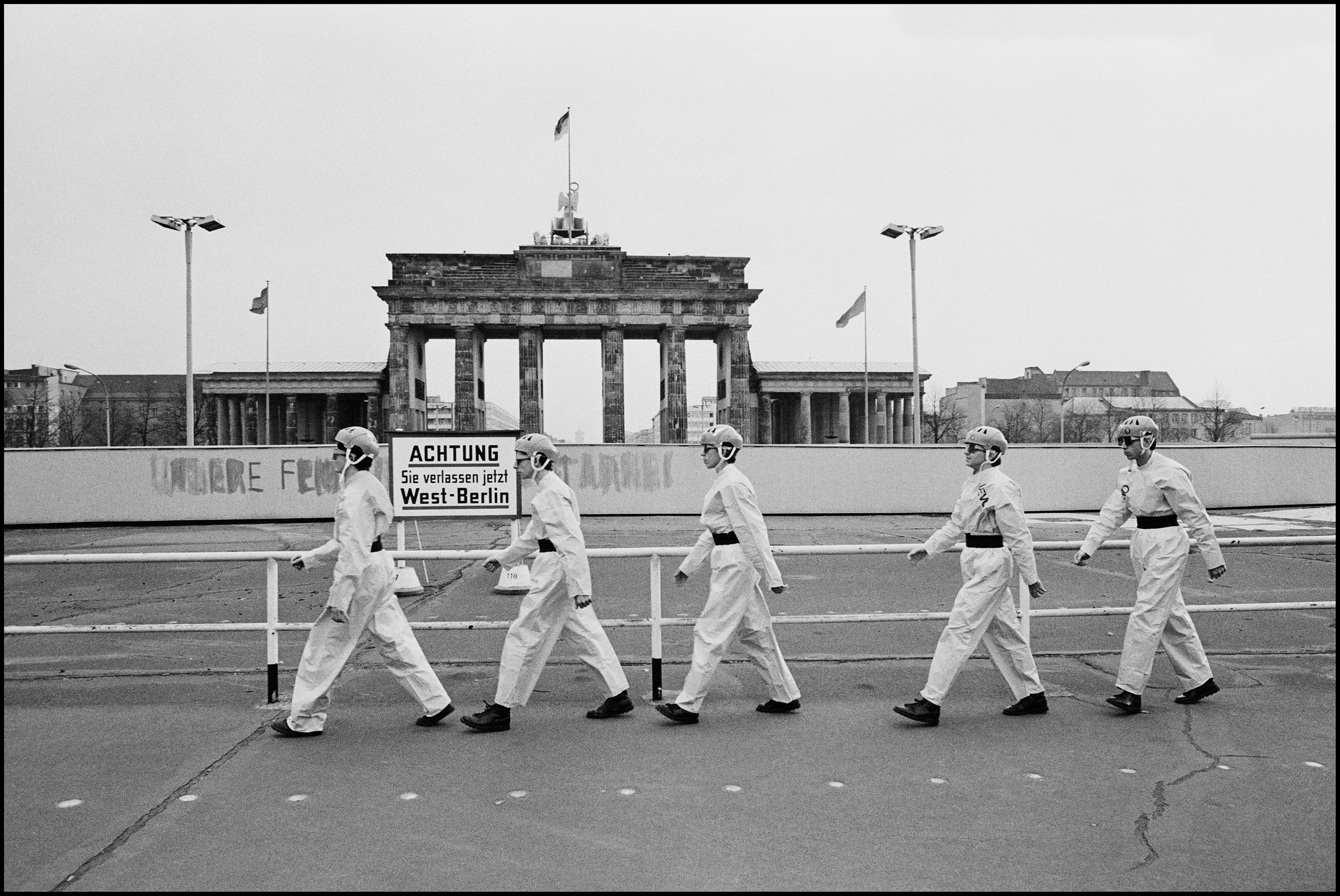 Dev at the Brandenburg Gate 1978 I went over to Berlin to photograph Devo, we did all the normal photo shoot stuff in the afternoon before they had to go to the Soundcheck. But it still said nothing other than, here are a bunch of weird Americans from Akron, Ohio wearing yellow paper suits and funny hats. I did have a better idea, but it would take a lot of persuasion to achieve it. They had to leave for the airport at seven am, I suggested leaving an hour early and going to the Brandenburg Gate to do photos. Berlin was still a divided city and the Wall separated east from west. I ran it by Gerry, I ran it by Mark, once I had their votes the rest of the band agreed, and so off we went. When we reached the Wall it was completely deserted, the band changed into their outfits on the coach while I checked out the best position for photographs. Then the band quickly posed together for a few frames, but it still didn't say that much about the situation. I asked them to march back and forth in front of the gate a few times. It was to be my East German homage to Abbey Road. Out of the corner of my eye I spotted some Russians on top of the gate, with what I assumed were binoculars. Just as the band crossed for the third time the Russians stood up and I saw it was not binoculars they were carrying it was machine guns. Then a bunch of German came out of the loudspeakers, it scared the life out of us, in fact the frames are not level because I was seriously spooked by the situation. They scarpered quickly back to the bus, I joined them and in moments we were soon on our way out of the country, the adrenaline rush was pretty strong, but, I doubt if the whole thing lasted more than three minutes, machine guns have that effect on people.