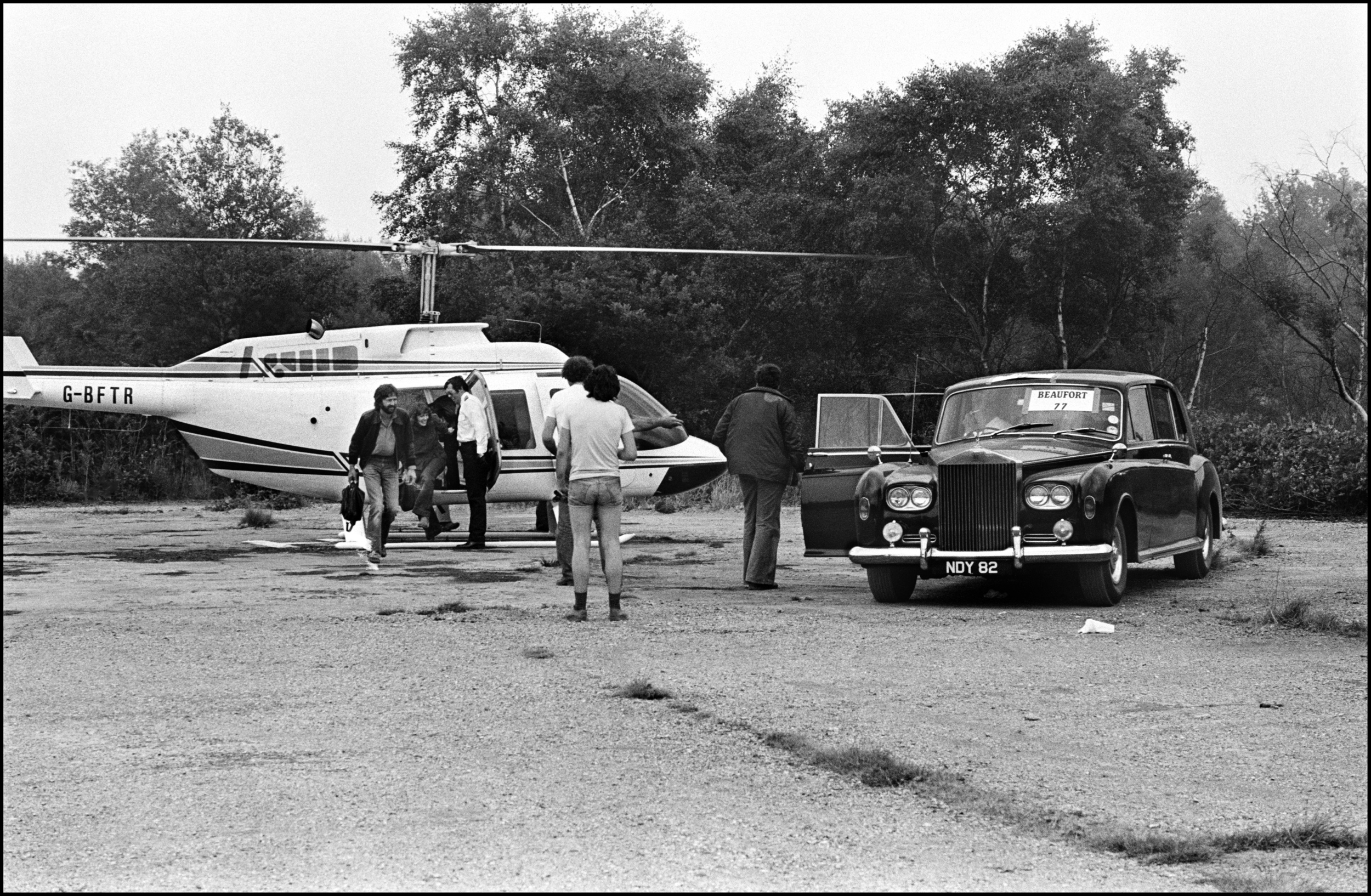 Eric Clapton Arrives at Blackbushe 1978 Here is a photograph that shows the level to which Rock Stars are pampered. It is taken Backstage at Blackbushe Airport at the Bob Dylan Concert. Dylan was quite happy to arrive in a coach with his Band, he enjoyed looking out if the window at the surrounding British countryside, but Eric Clapton had a different idea, he wanted to excerpt the minimum effort to get to the gig, and wanted it done in the shortest time possible. So a Helicopter was sent to his Country House where it landed in on his lawn. Eric walked the short distance from his house to the waiting Helicopter. After a journey of less than thirty minutes he arrived at the Gig, the Helicopter landed about 200 yards from the Backstage area, but to save him from any physical exertion and to keep him in the luxurious style he was used to a Rolls Royce was parked nearby. Eric then walked over to the waiting Rolls, got into the back seat, and was driven to his dressing room, a journey which took less than a minute to complete. How nice it must be to live inside a superstar bubble, to have everything at your disposal, to simply have to walk about twelve steps from Helicopter to Rolls Royce. But, to me, as well as documenting one of Rock's more surreal moments, it shows off the impeccable style of the British Roadie, just take a look at the guy's shorts, it's living proof of why Roadies often got more girls than the Band.