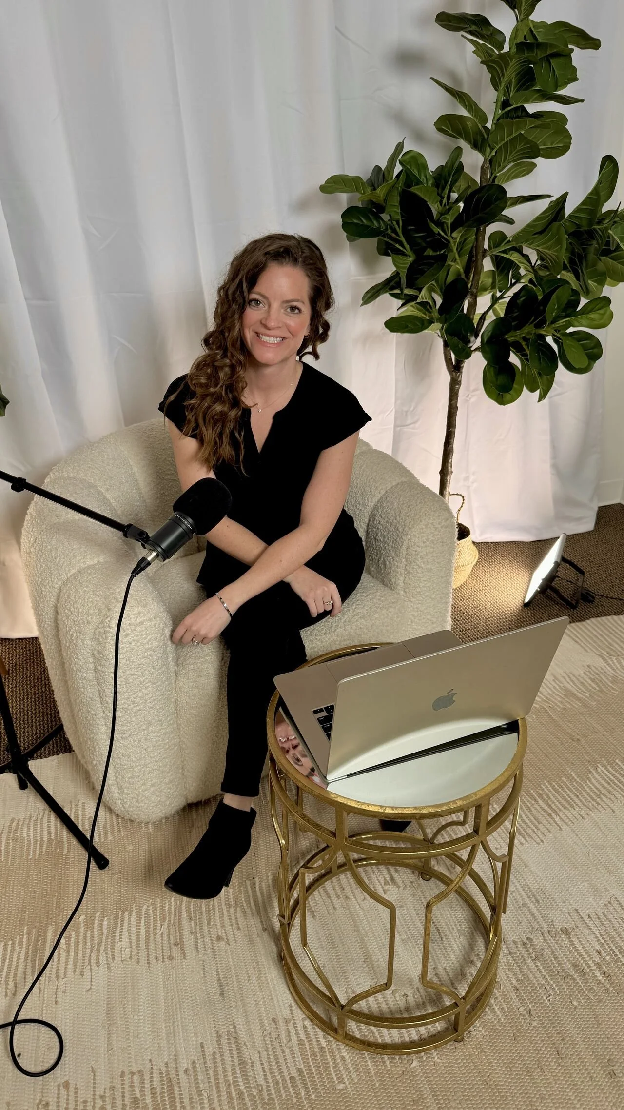 A woman with long, curly brown hair sitting on a cream-colored textured armchair. She is smiling, wearing a black top and black pants, and has a microphone on a stand positioned near her mouth. A silver MacBook laptop is open on a small gold table wi