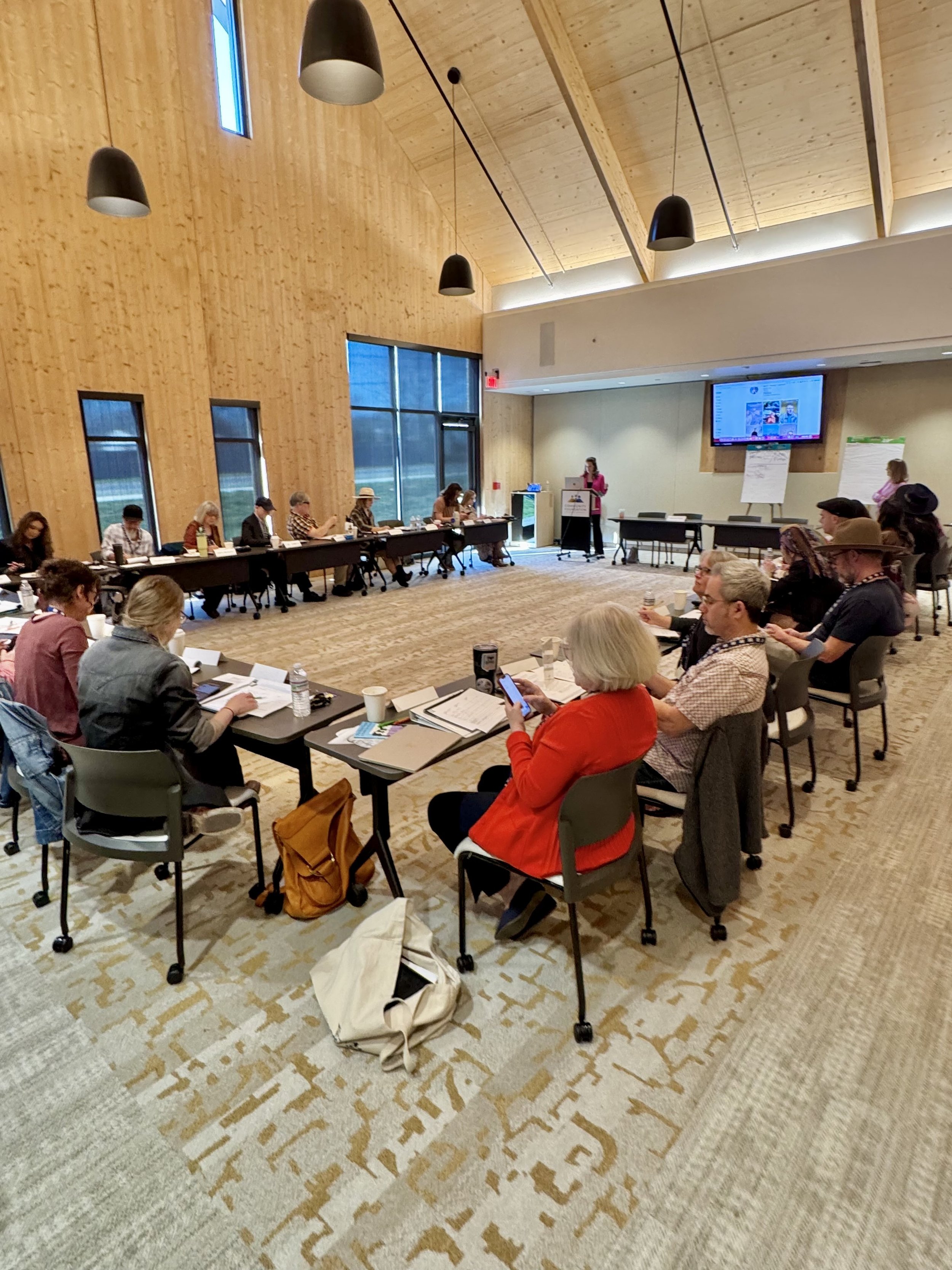 A large conference room filled with people seated around a U-shaped arrangement of tables. A woman is presenting at a podium in front of a large screen. Attendees are engaged, some taking notes or using phones. The room has high wooden ceilings, larg