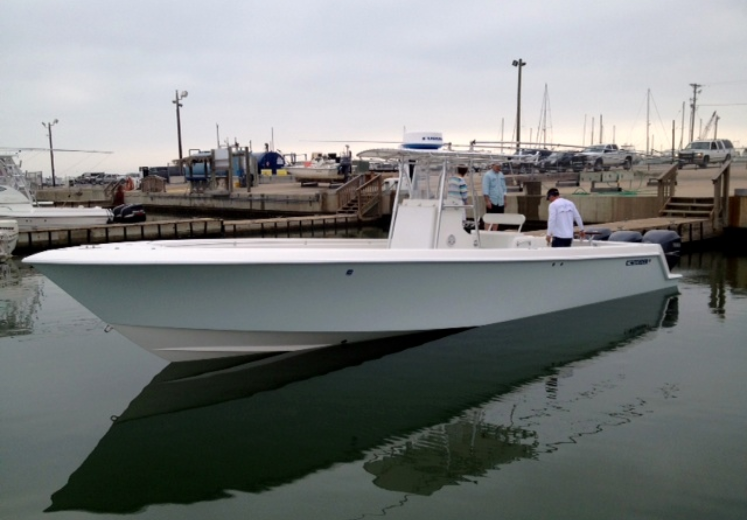 A white boat docked at a marina with several people on board and on the dock, and boats and cars in the background.