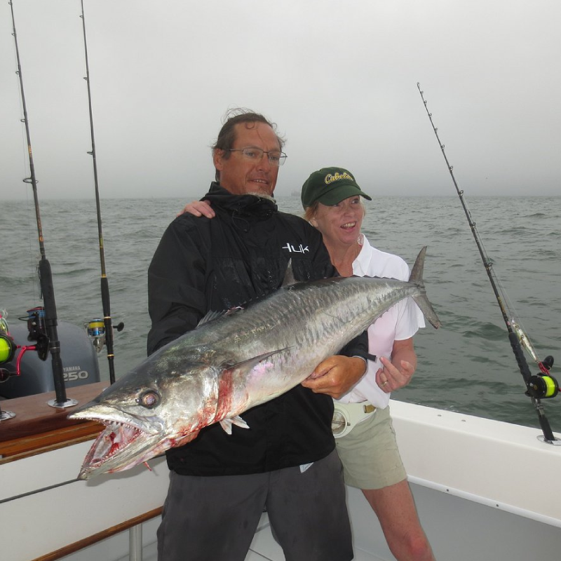 Two people on a boat holding a large fish, fishing rods in the background, on a cloudy day at sea.