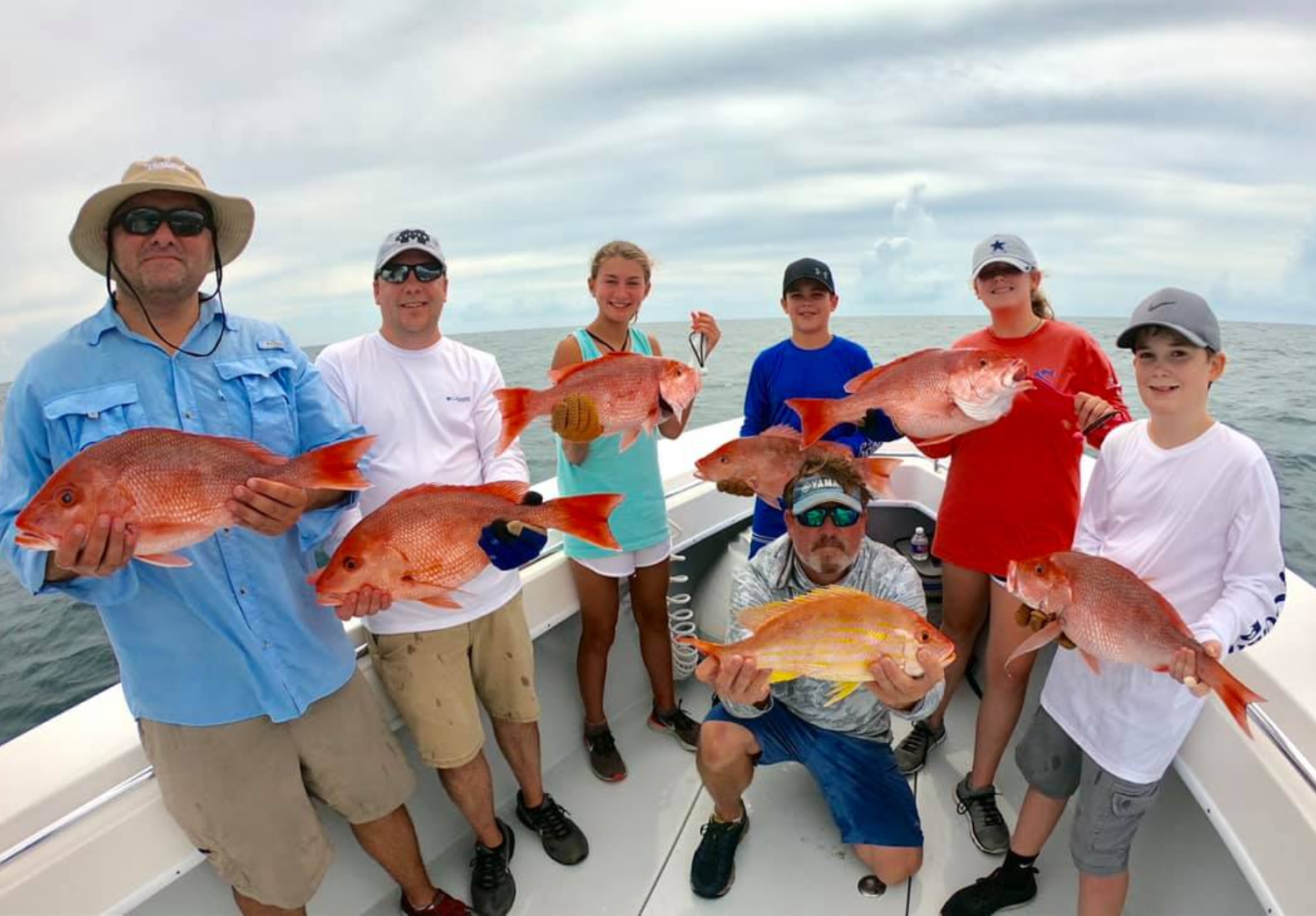 Group of people on a boat holding large red snapper fish, with a cloudy sky and ocean in the background.