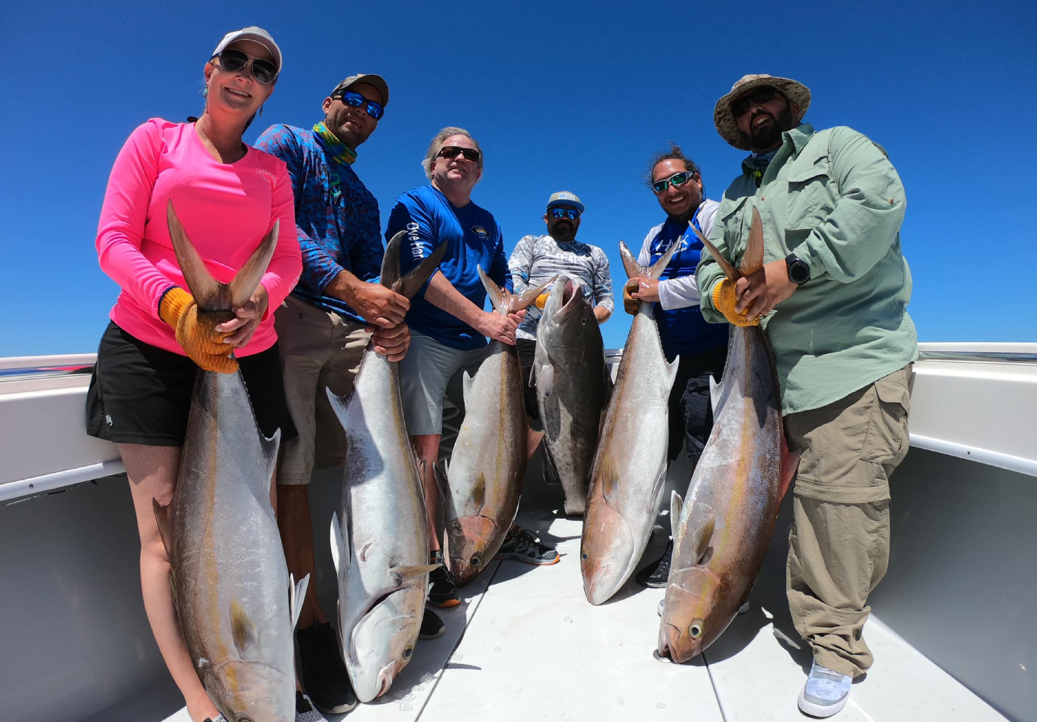 Group of six people on a boat holding large fish they caught during a fishing trip, with a clear blue sky in the background.