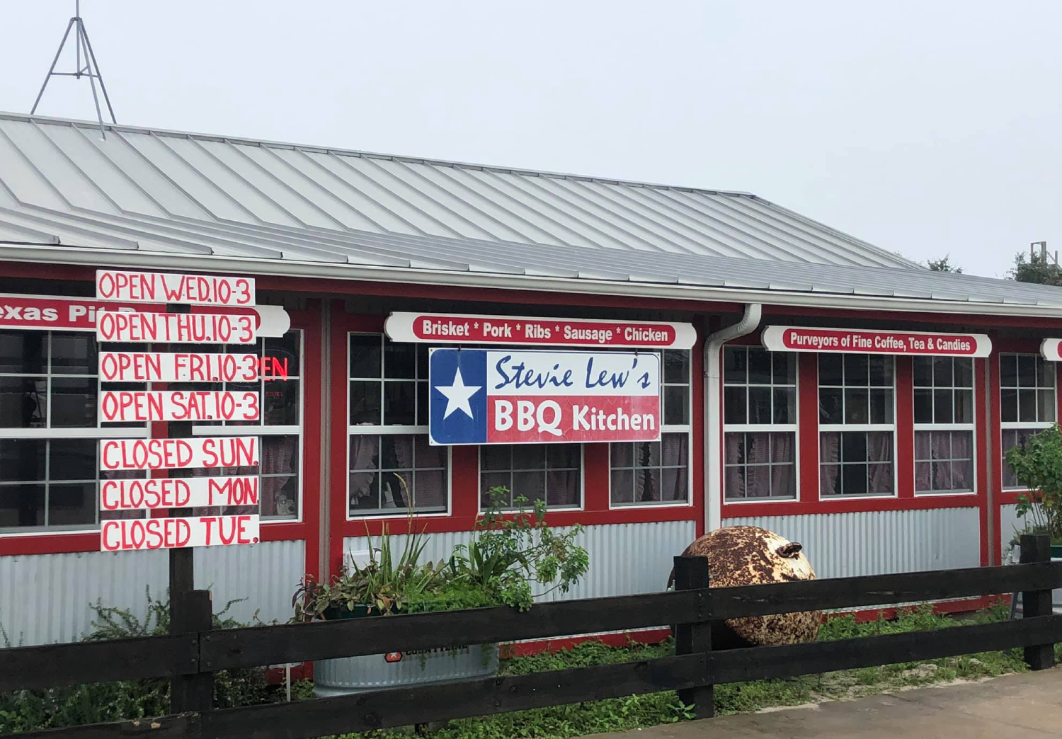 Exterior of Stevie Lew's BBQ Kitchen with signs displaying operating hours and menu items, including brisket, pork, ribs, sausage, chicken, coffee, tea, and candies, and a large rusty metal object in the foreground.