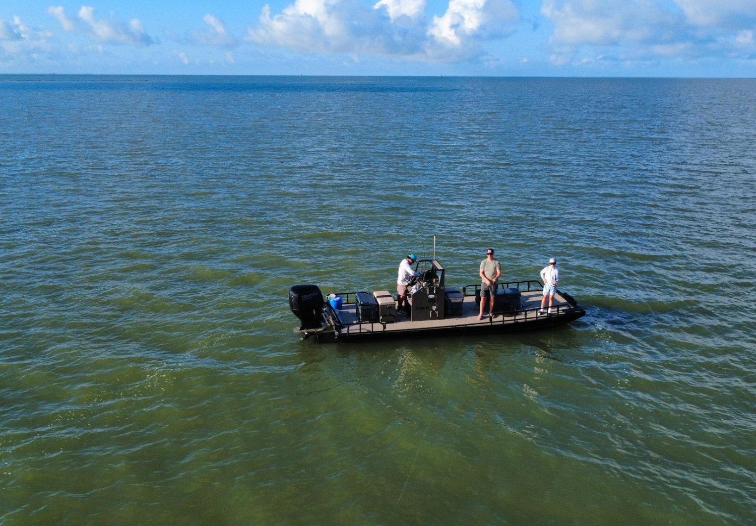 Three people on a small black boat floating on water with greenish tint, under a partly cloudy sky.