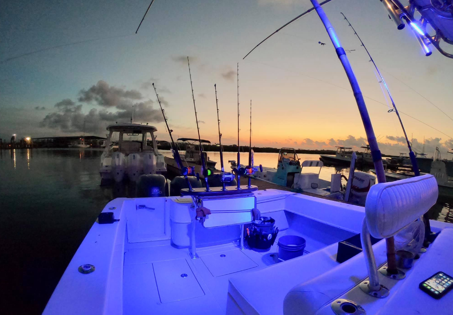 View of boats docked at a marina during sunset, with fishing rods mounted on a boat in the foreground.
