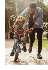 Photograph of father running alongside young boy pedalling bike as he is learning to ride.