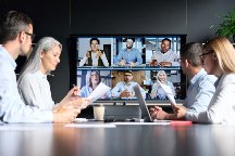 Two male and two female professionals seated on either side of a board room table, facing a television mounted at the end of the room and talking with six other team members on a video call.
