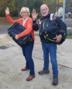 Sue and Paul Ebbers waving to the reader, with their luggage bags in hand, together facing left and smiling about their upcoming vacation.