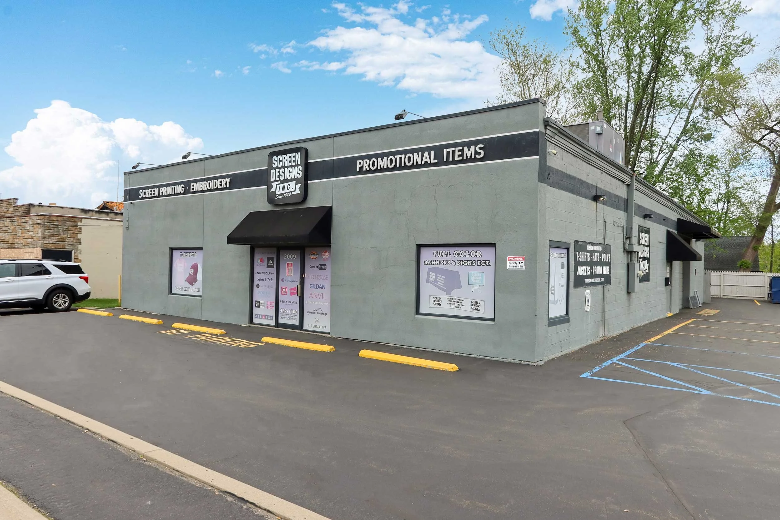 A commercial building with signs for screen printing, embroidery, promotional items, and screen design services. The building has a gray exterior with black awnings over the windows and entrance. There is a parking lot with yellow bumpers and a silver SUV parked on the left side, with some trees and a clear sky in the background.