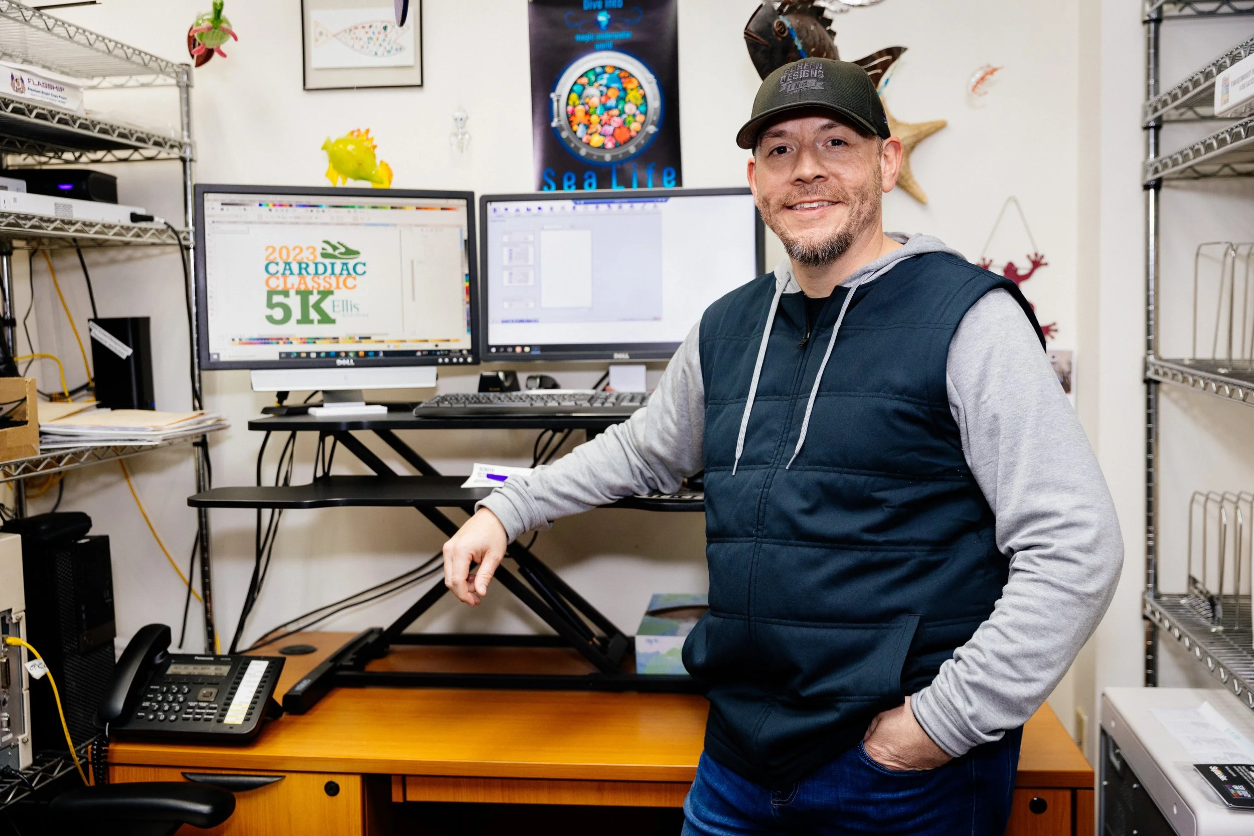 Man standing in an office with dual computer monitors, one displaying a colorful graphic with '2023 Cardiac Classic 5K,' and the other showing design software. There are wire shelving units, a landline phone, and various office supplies around him.