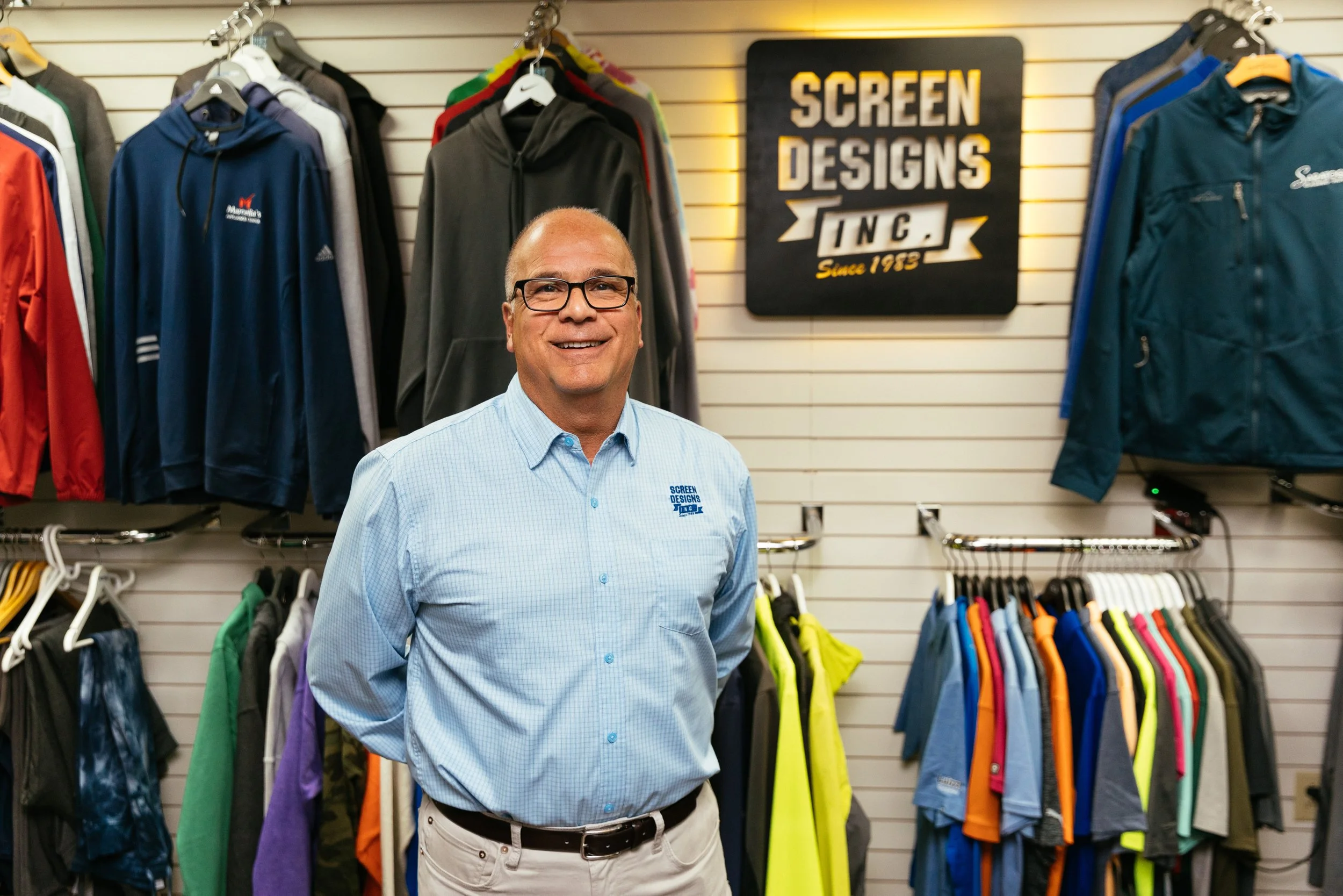 A man in a light blue checkered shirt with glasses standing in front of a display of colorful jackets and hoodies in a store, with a sign that reads "Screen Designs Inc. Since 1983."
