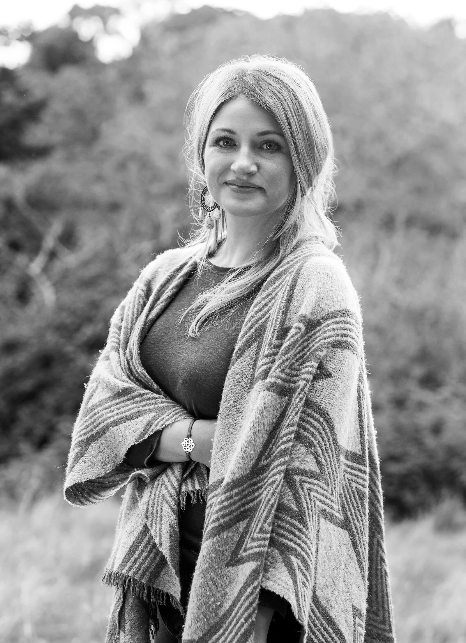 A woman standing outdoors on a blurred natural background, wearing a patterned shawl and a dark top, with her arms crossed and looking at the camera.