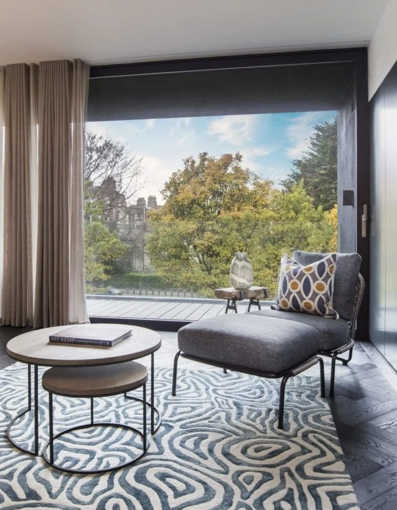 A modern living room with a large window overlooking trees with autumn leaves, a dark armchair with a patterned pillow, a small side table with a decorative vase, a round coffee table with a book on it, and a patterned rug.