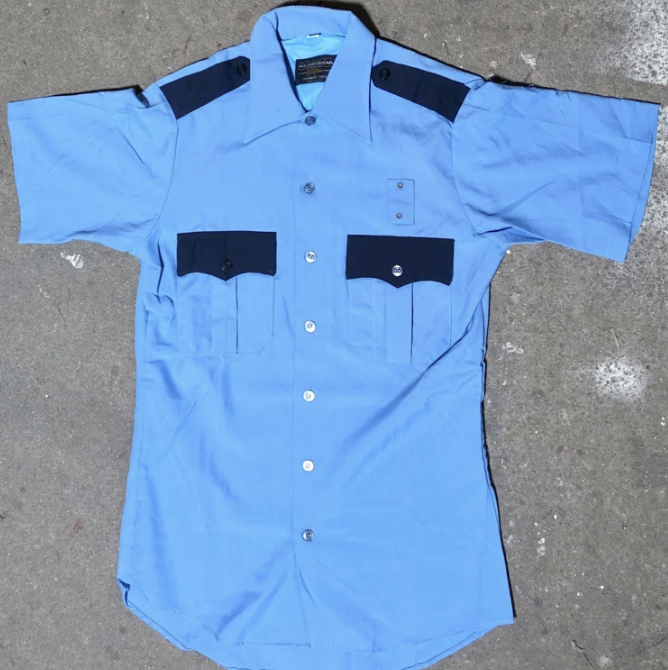 Light blue police or security shirt with dark blue shoulder patches and chest pockets, laid flat on a concrete surface.