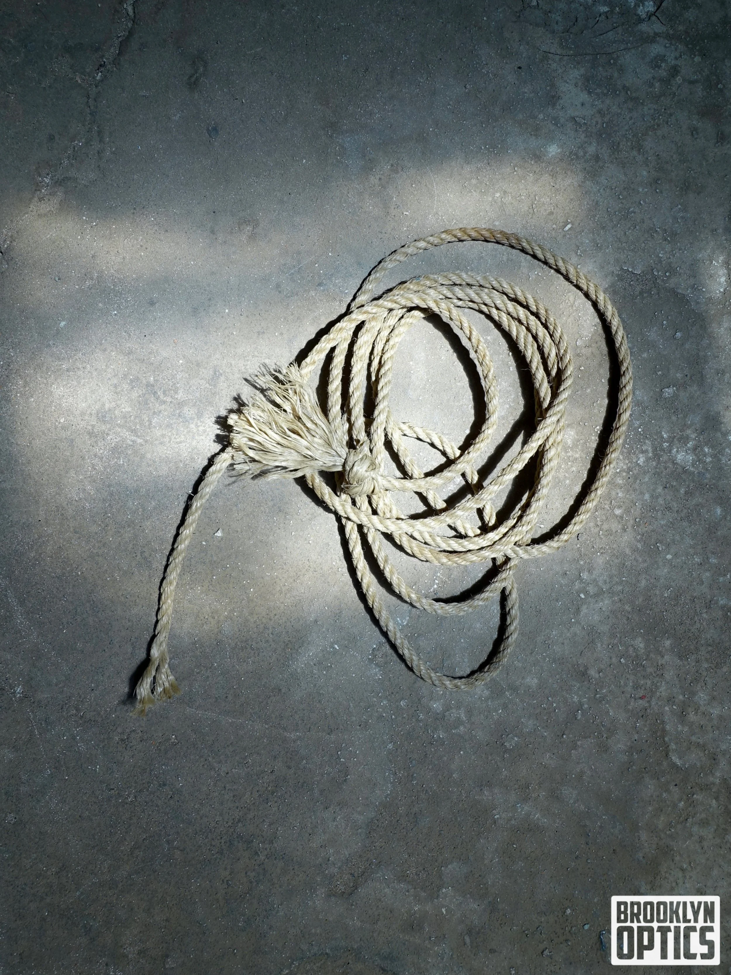 A tangled piece of beige rope lying on a concrete floor, with the logo 'Brooklyn Optics' in the bottom right corner.