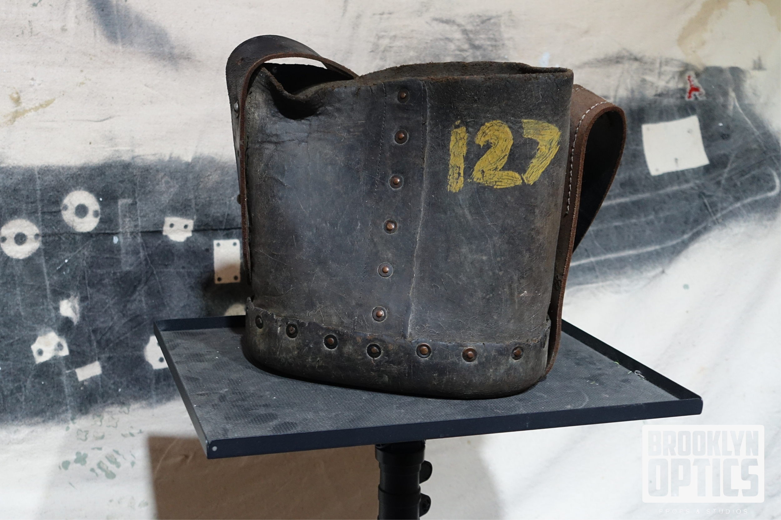 Old, worn black leather military-style bag with yellow numbers '127' painted on the front, sitting on a black metal table.