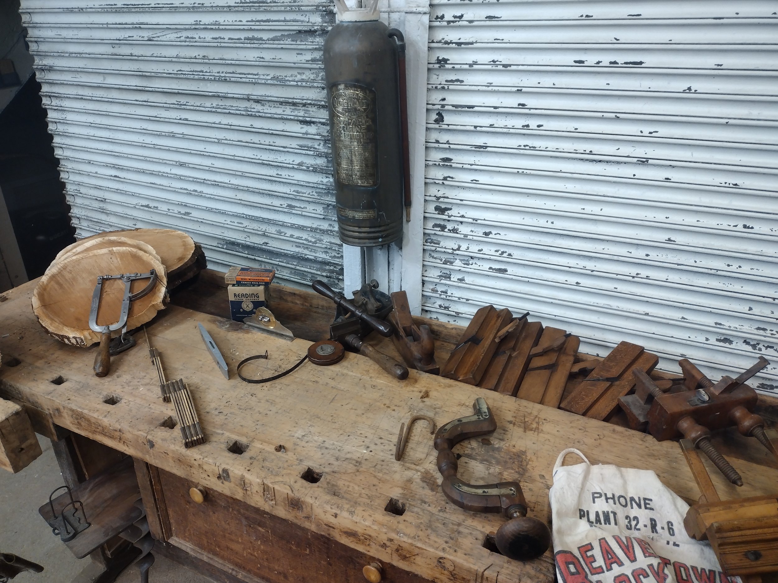 A woodworking workbench with a slice of wood, C-clamp, sanding paper, and various hand tools in a workshop with a white, weathered metal wall.