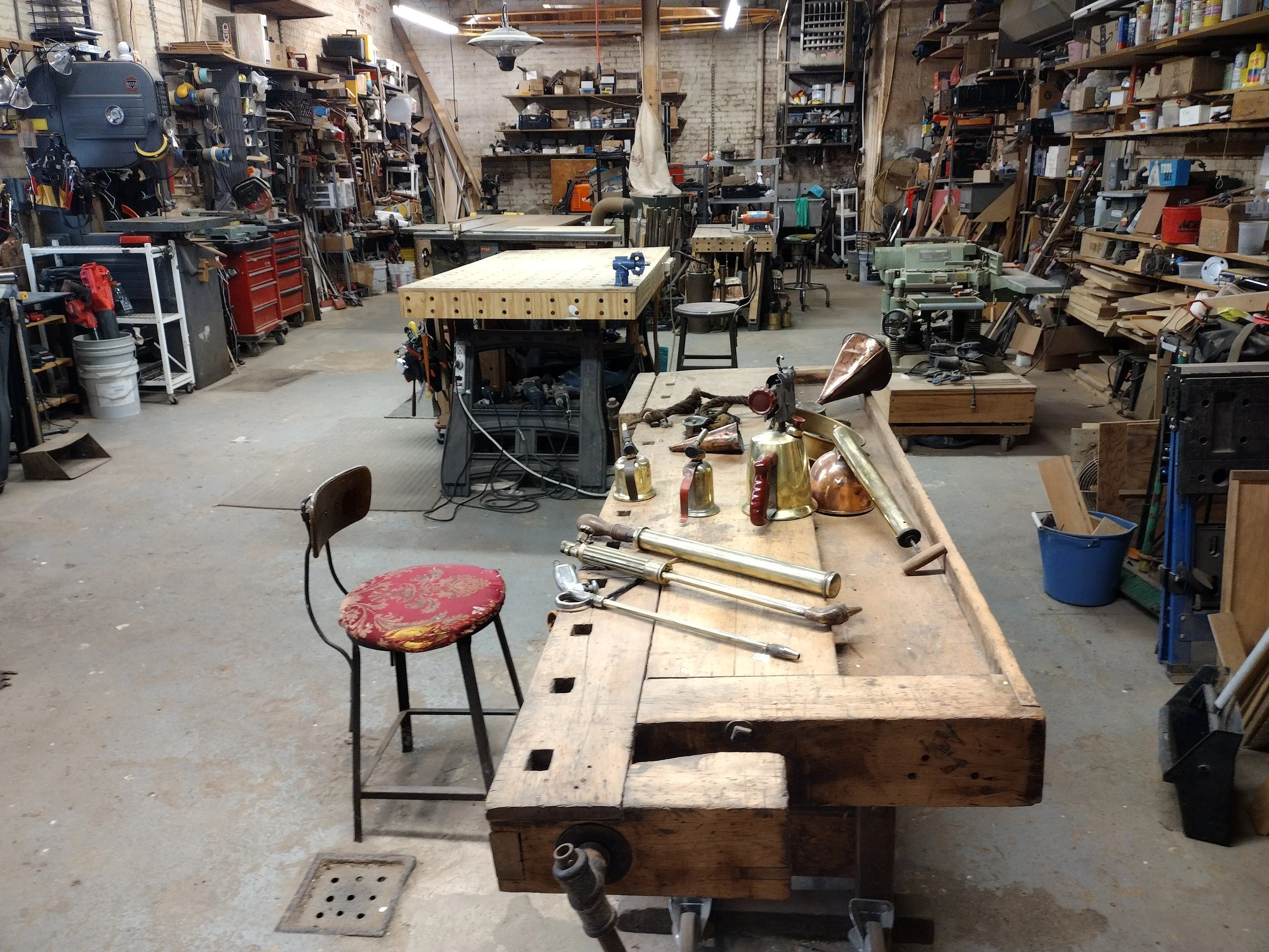 Workshop with a wooden workbench holding vintage brass and metal objects, tools, and pipes. Shelves filled with various equipment and tools line the walls, plus a chair with a patterned cushion in the foreground.