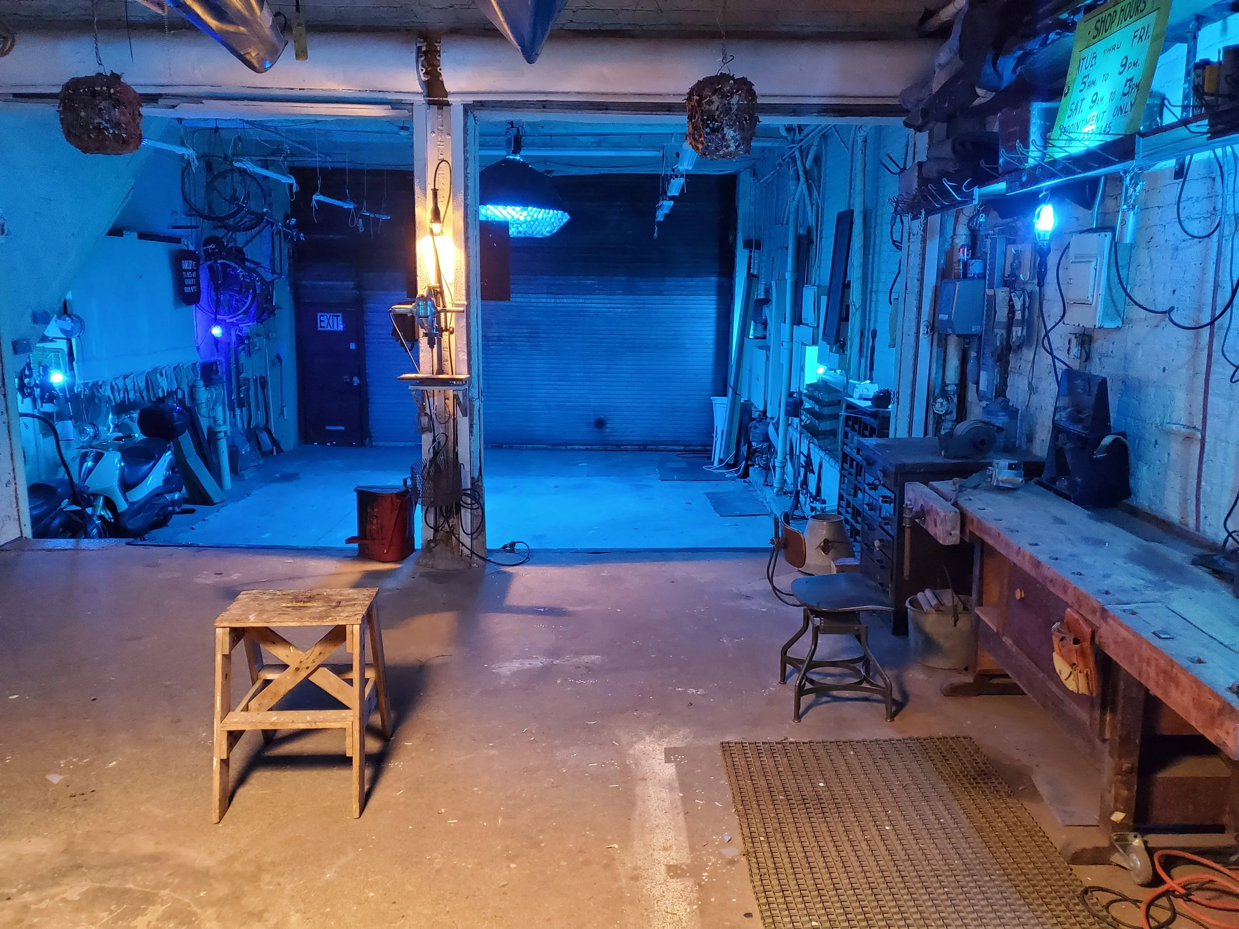 Interior of a workshop or garage with tools hanging on the walls and workbenches, illuminated by blue and yellow lighting, with a partially open garage door at the back.