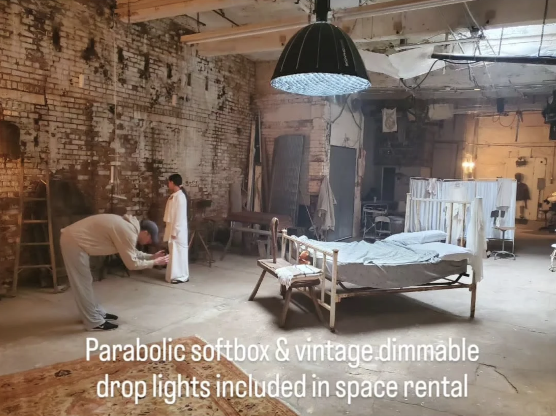 An old, rustic room with a brick wall, a hospital bed with a vintage or antique frame, and various furniture and equipment. Two men are adjusting a piece of equipment, and there is a large black parabolic softbox light hanging from the ceiling. The space has a gritty, industrial look, and it appears to be set up for a photo or film shoot, with a caption indicating that the space rental includes a parabolic softbox and vintage dimmable drop lights.