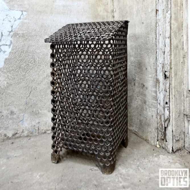 A vintage metal wine rack with circular holes, standing on four small legs against a weathered wall.