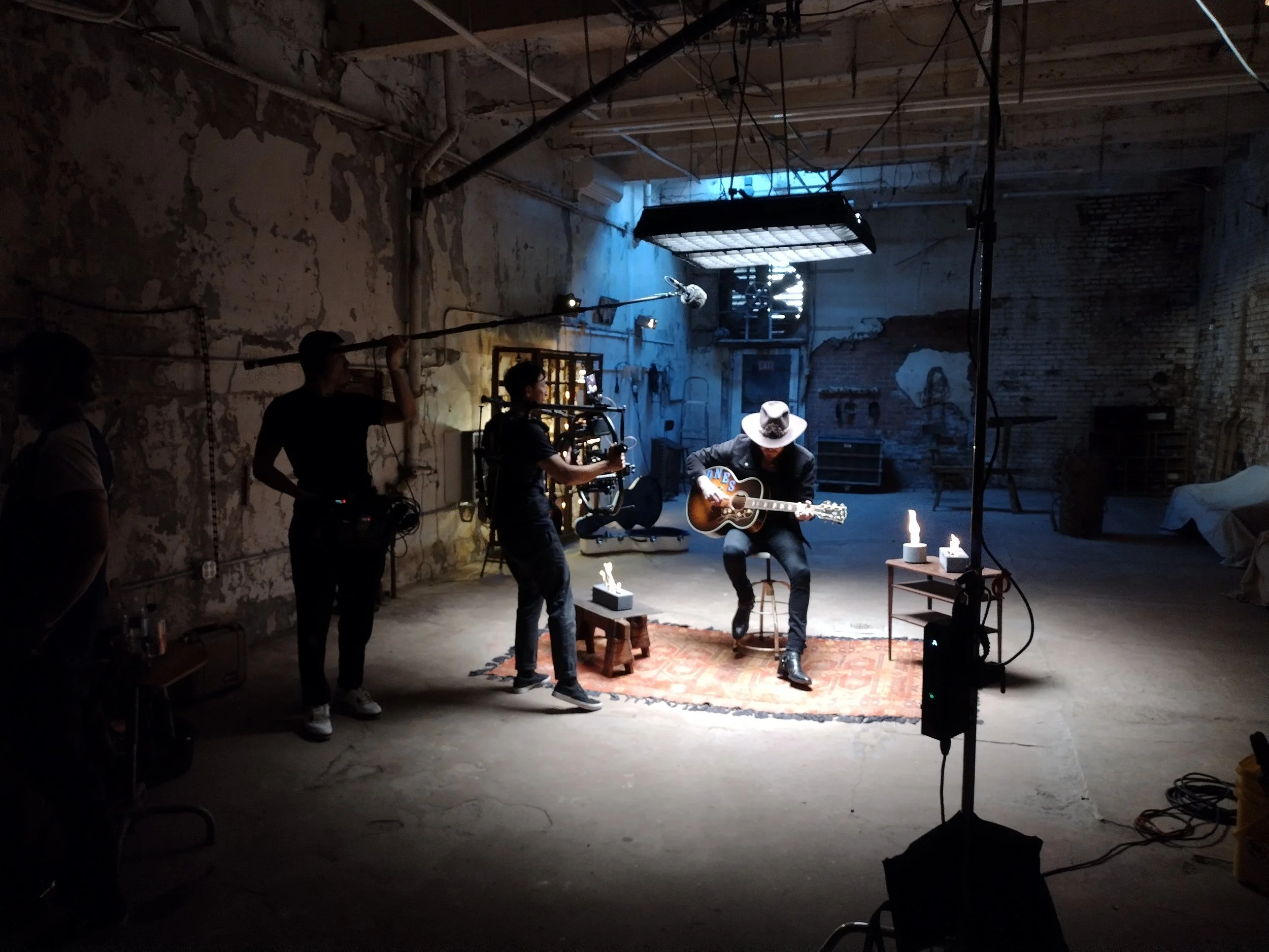 Behind-the-scenes of a music video or photo shoot in an industrial space with a person playing guitar, surrounded by crew members and filming equipment.