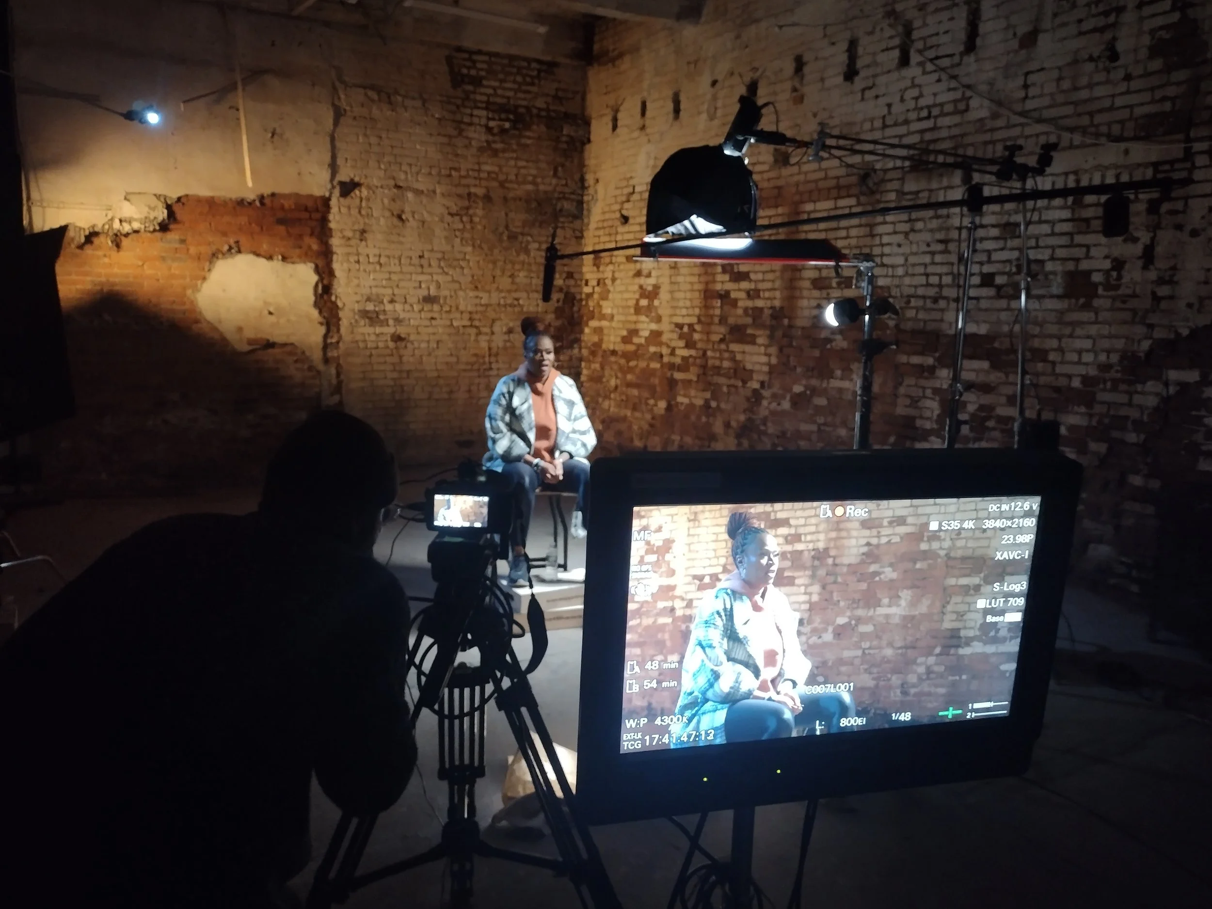A woman sits on a chair in a studio with exposed brick walls, being recorded on video with professional lighting and camera equipment.