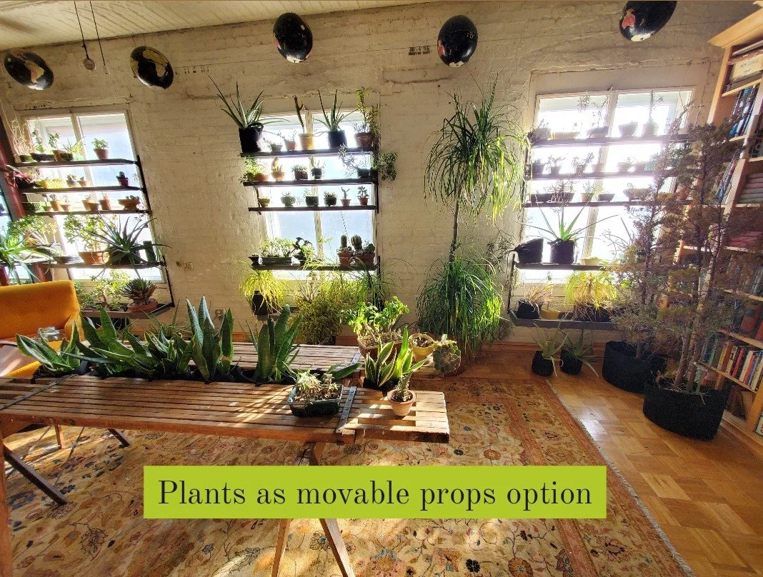 A cozy room filled with various houseplants placed on shelves, tables, and the floor, with sunlight coming through large windows. The room has wooden floors, a brick wall, and a bookshelf on the right. The caption reads "Plants as movable props option."
