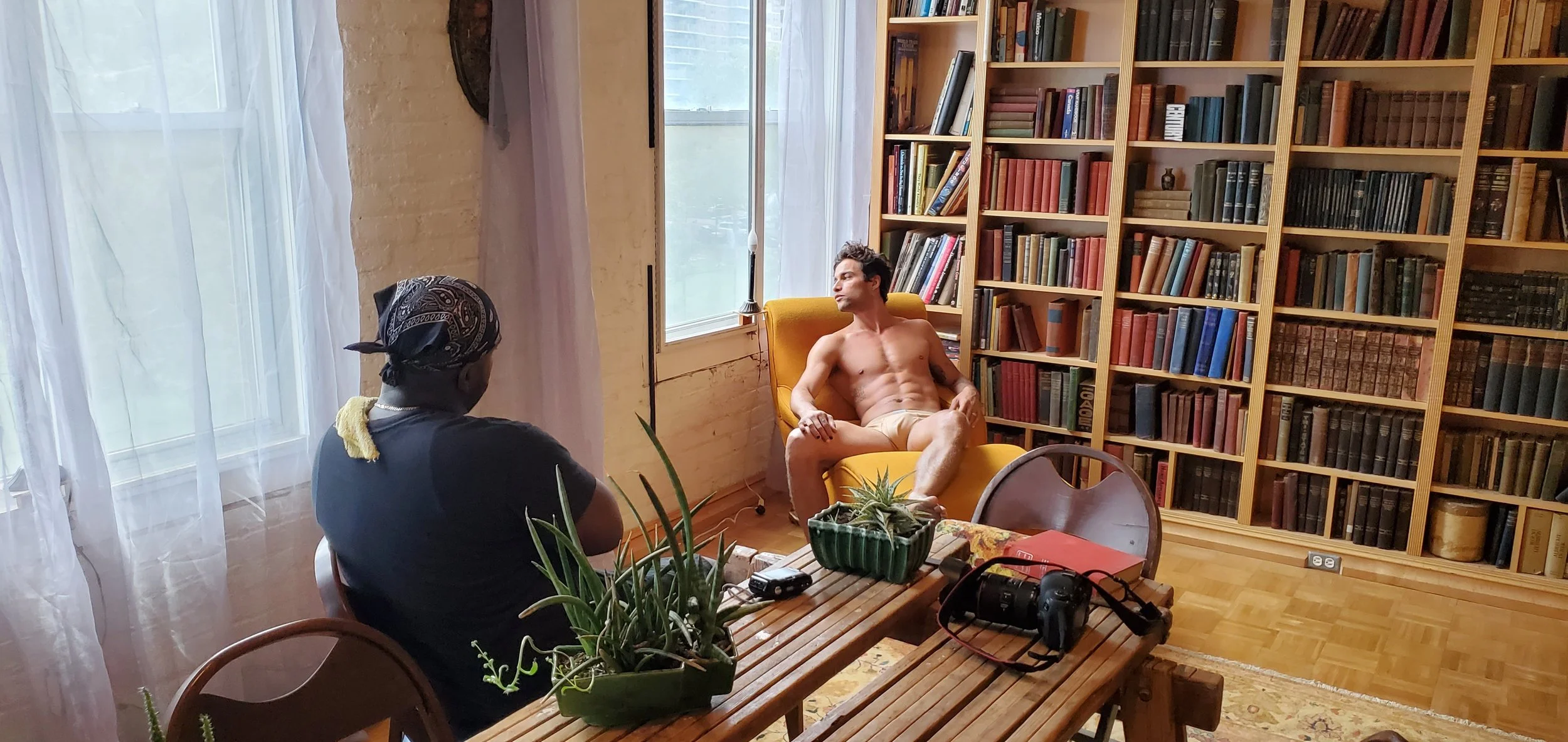 A shirtless man sitting in a yellow armchair by a window, with a woman sitting at a wooden table with plants and camera gear, in a room with bookshelves.