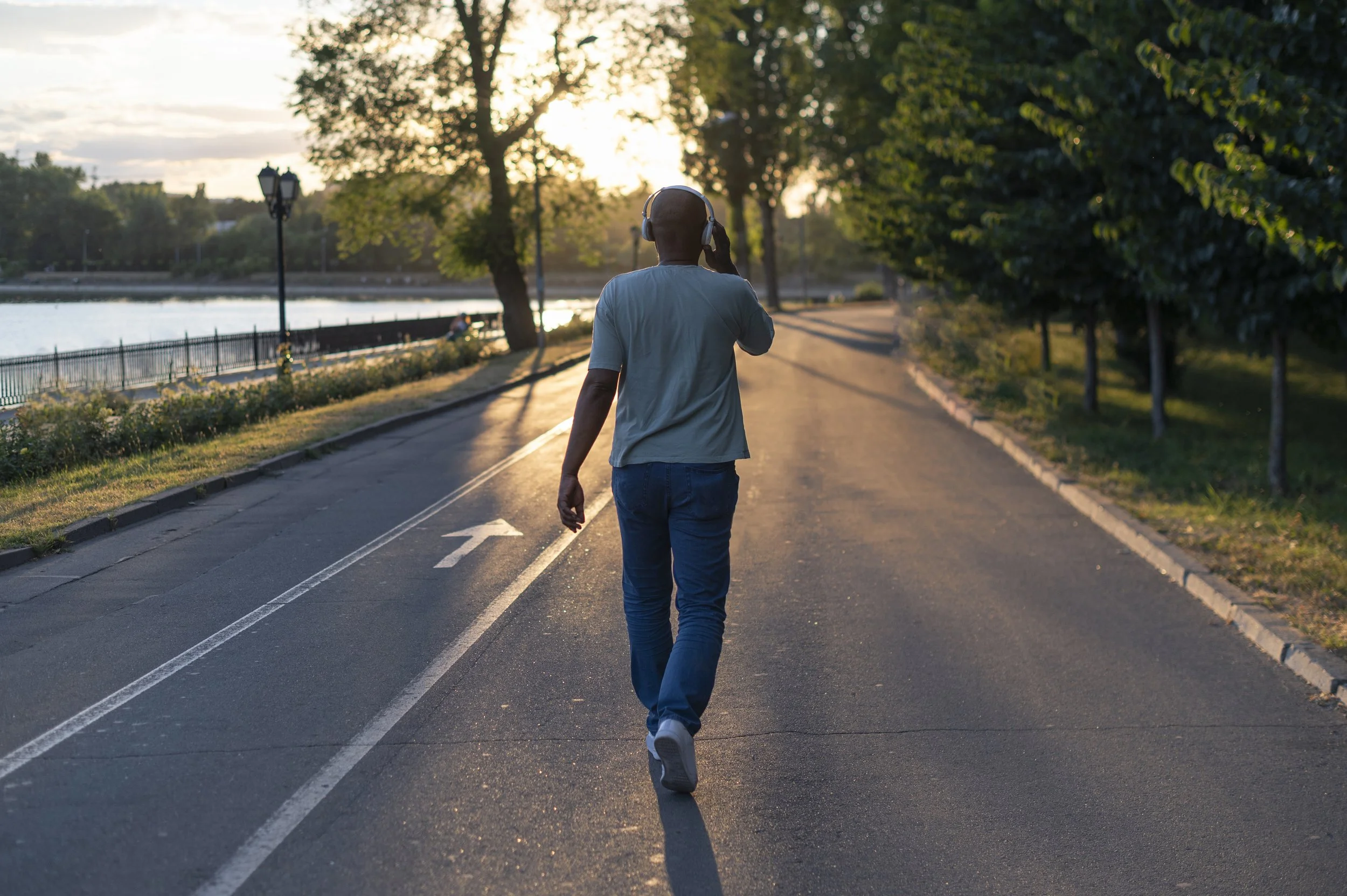 A black man is walking up a street with his back to us and headphones on. There is sun peaking through trees up ahead in the distance.
