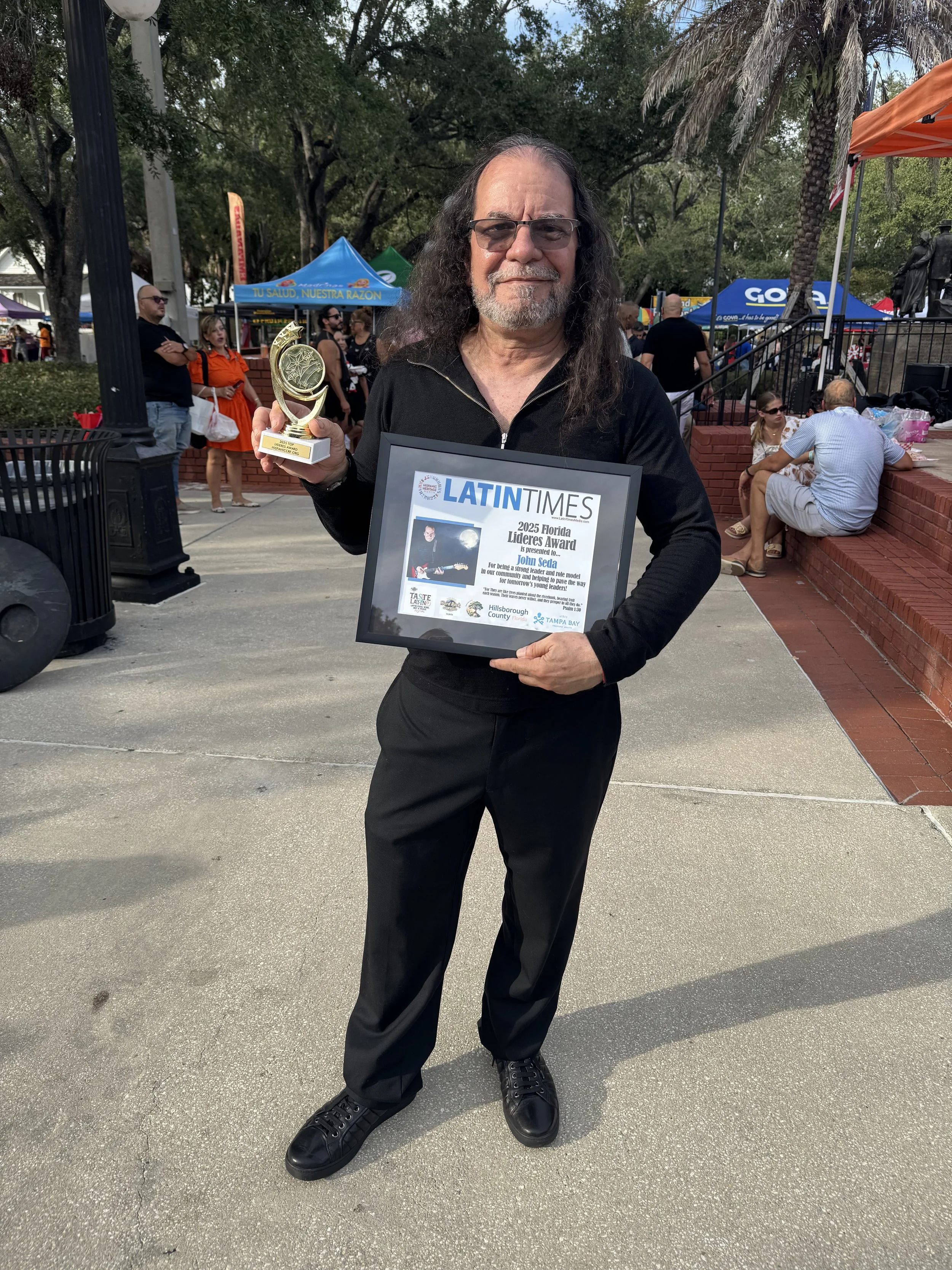 Music A man with long hair and glasses holding a trophy in his right hand and a framed certificate in his left in an outdoor park with tents and people in the background.