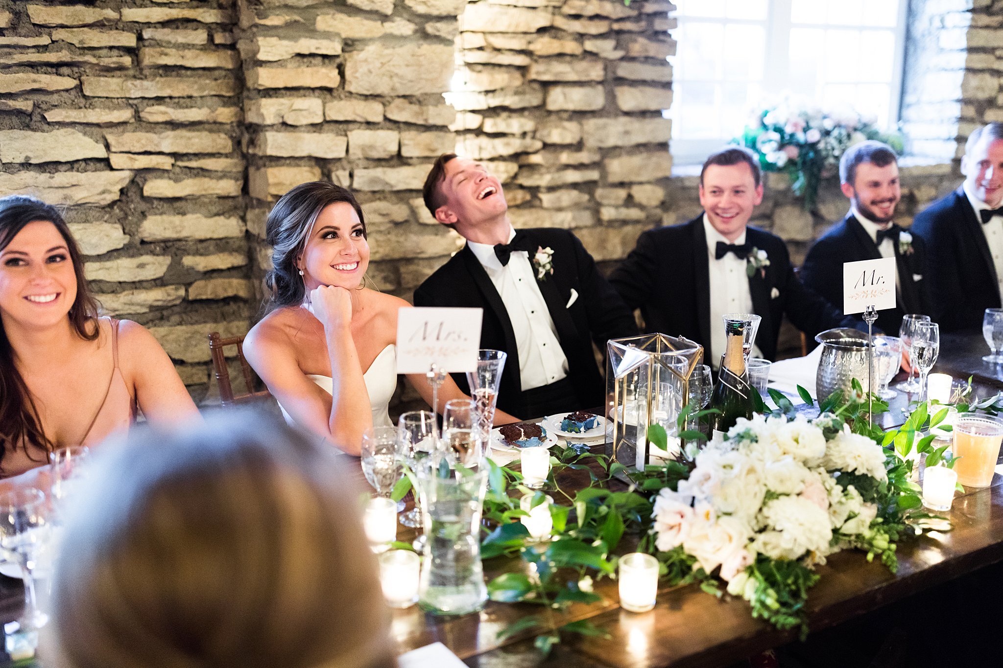  Bride and groom laughing while seated at head table during speeches. 