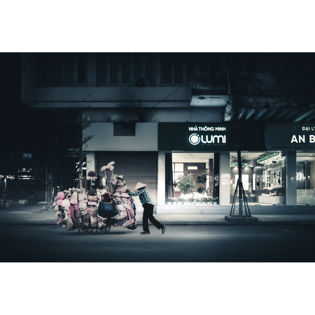 Night street scene in Ha Long Bay: Vietnamese vendor in conical hat pushes cart loaded with household goods and toys past illuminated shopfront.