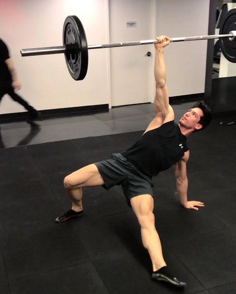 Chris Chang performing a Turkish get-up exercise with a barbell in a gym.