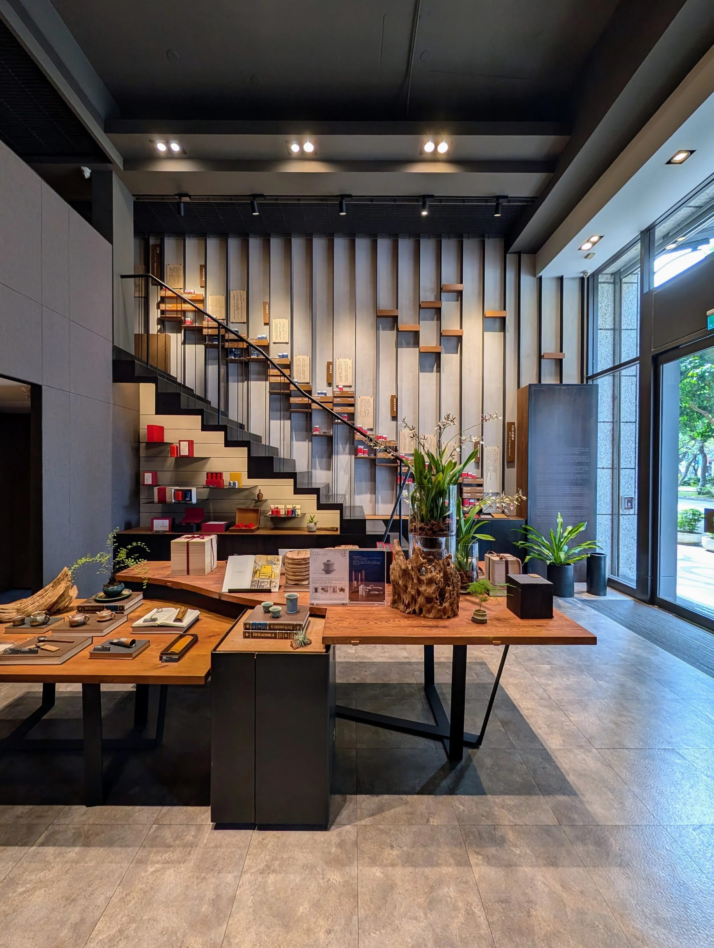 The interior of the tea store Wang De Chuan. Orchids and green plants, a table with teaware in the foreground and a staircase with a tea display wall in the background