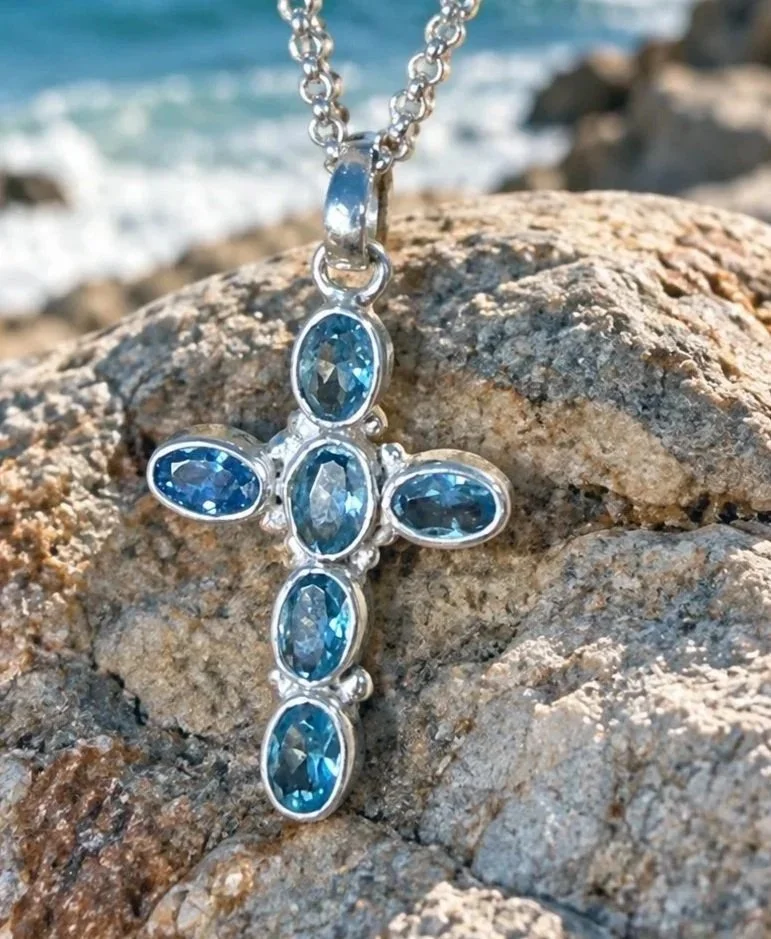 Blue Topaz Cross Pendant in sterling silver handmade in Bali displayed on a coastal rock