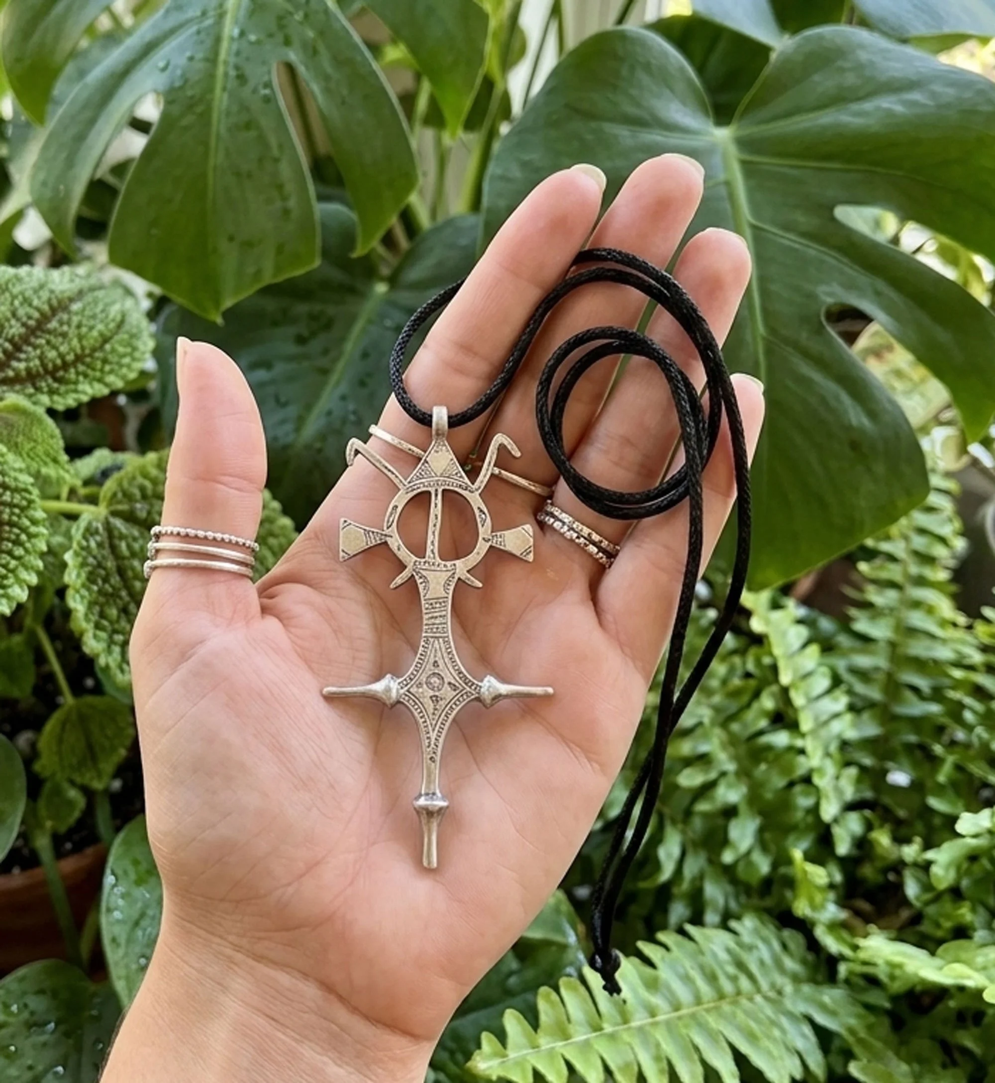 Hand holding a Moroccan Tuareg silver cross pendant necklace with intricate tribal detailing on a black cord surrounded by lush tropical greenery. Artisan boho statement jewelry.