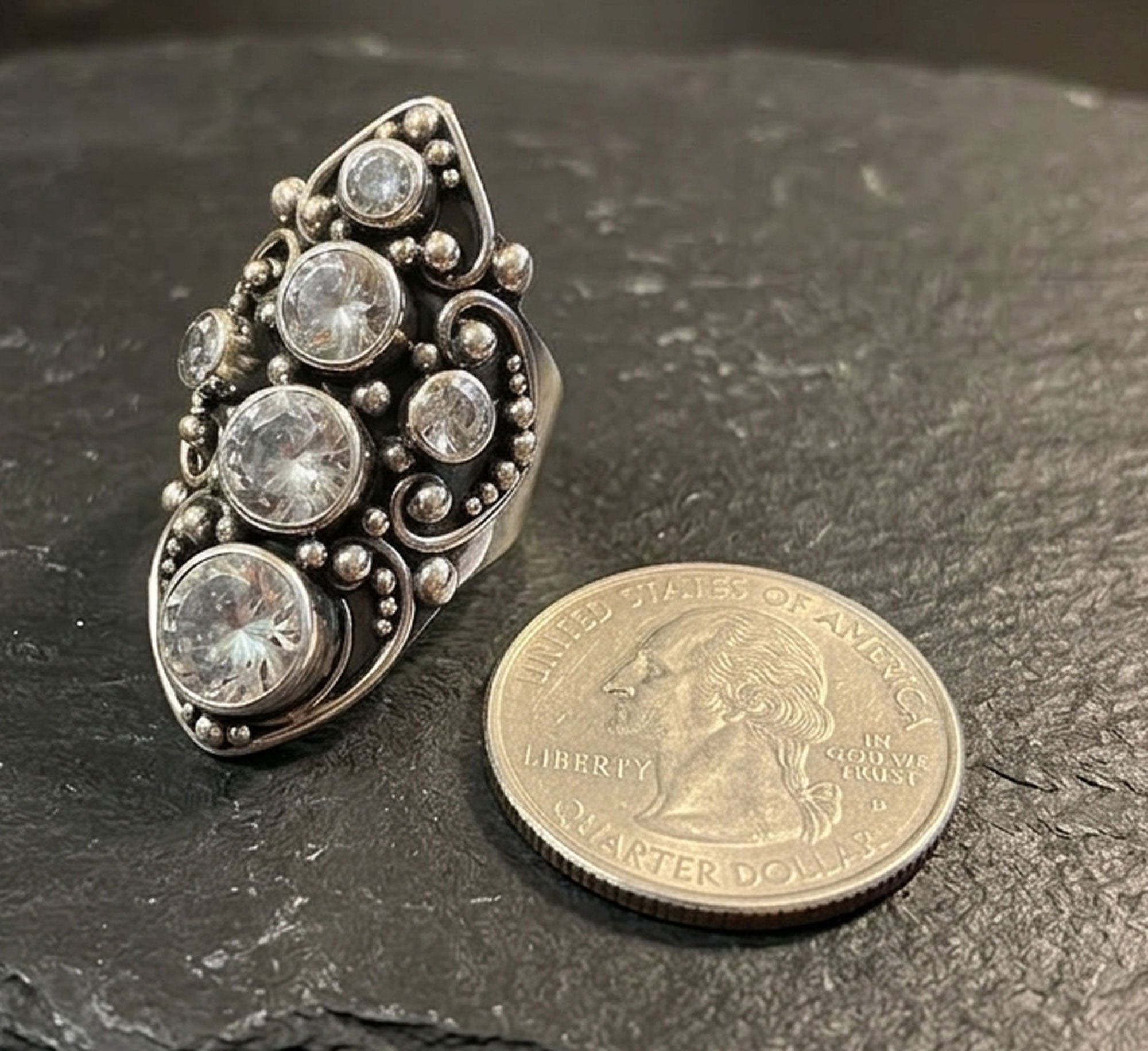 Close-up of handmade sterling silver cluster ring with large and small Zircon gems beside U.S. quarter on dark slate background.
