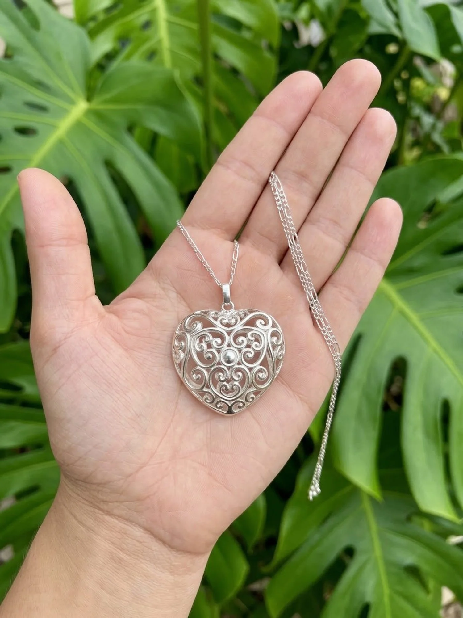 Large sterling silver openwork heart pendant displayed in a hand against green leaves