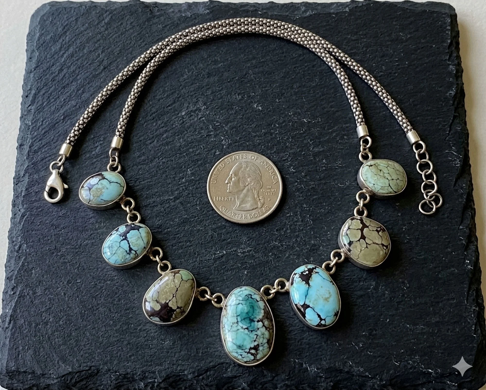Turquoise drop necklace in sterling silver with natural matrix stones displayed on slate with coin for size reference artisan jewelry