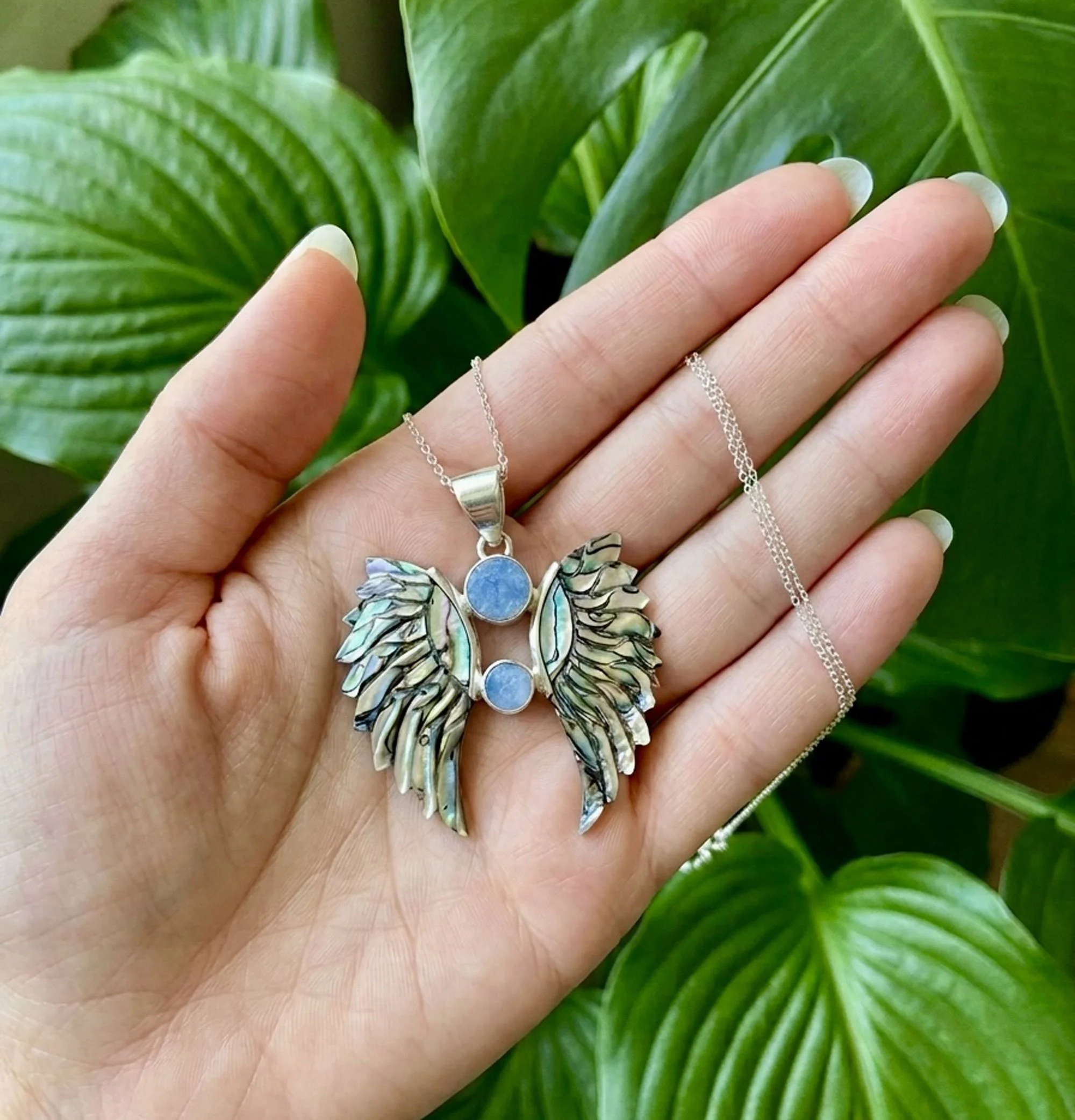 Handcrafted sterling silver angel wings pendant featuring genuine opal gemstones and iridescent abalone shell wings held in hand.