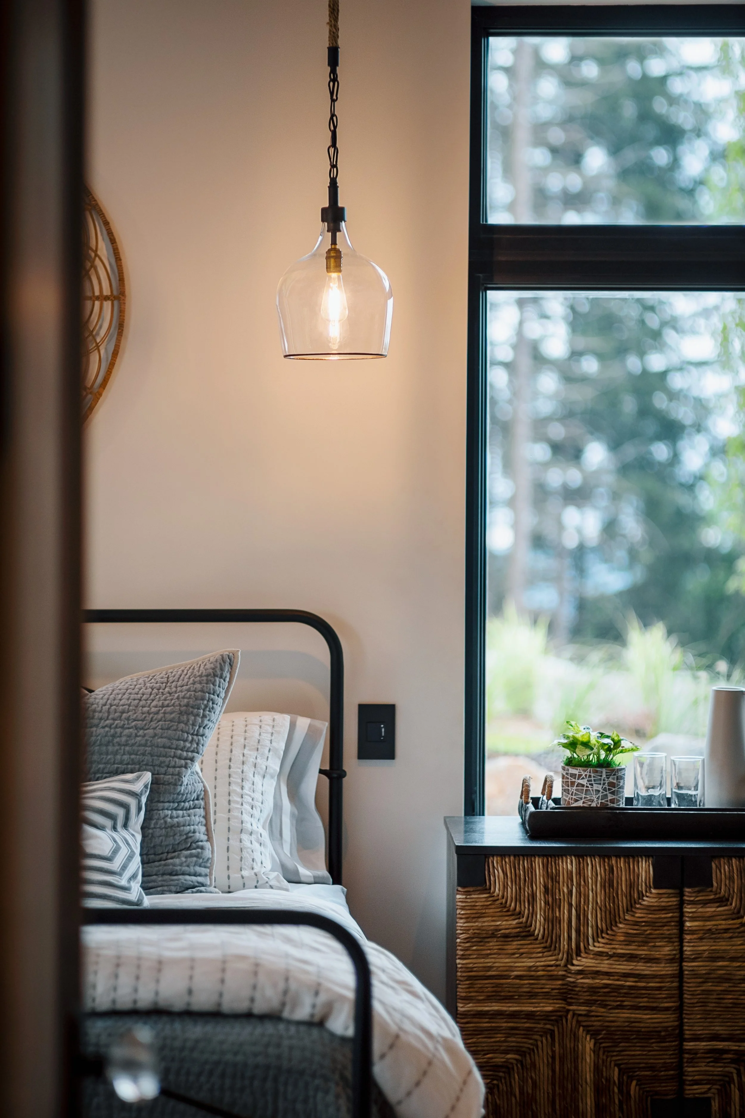 A bedroom with a black metal-framed bed, decorative pillows, and a wooden dresser. A glass pendant light hangs from the ceiling. Large window with a view of trees outside and a table with a plant, glassware, and a tray.