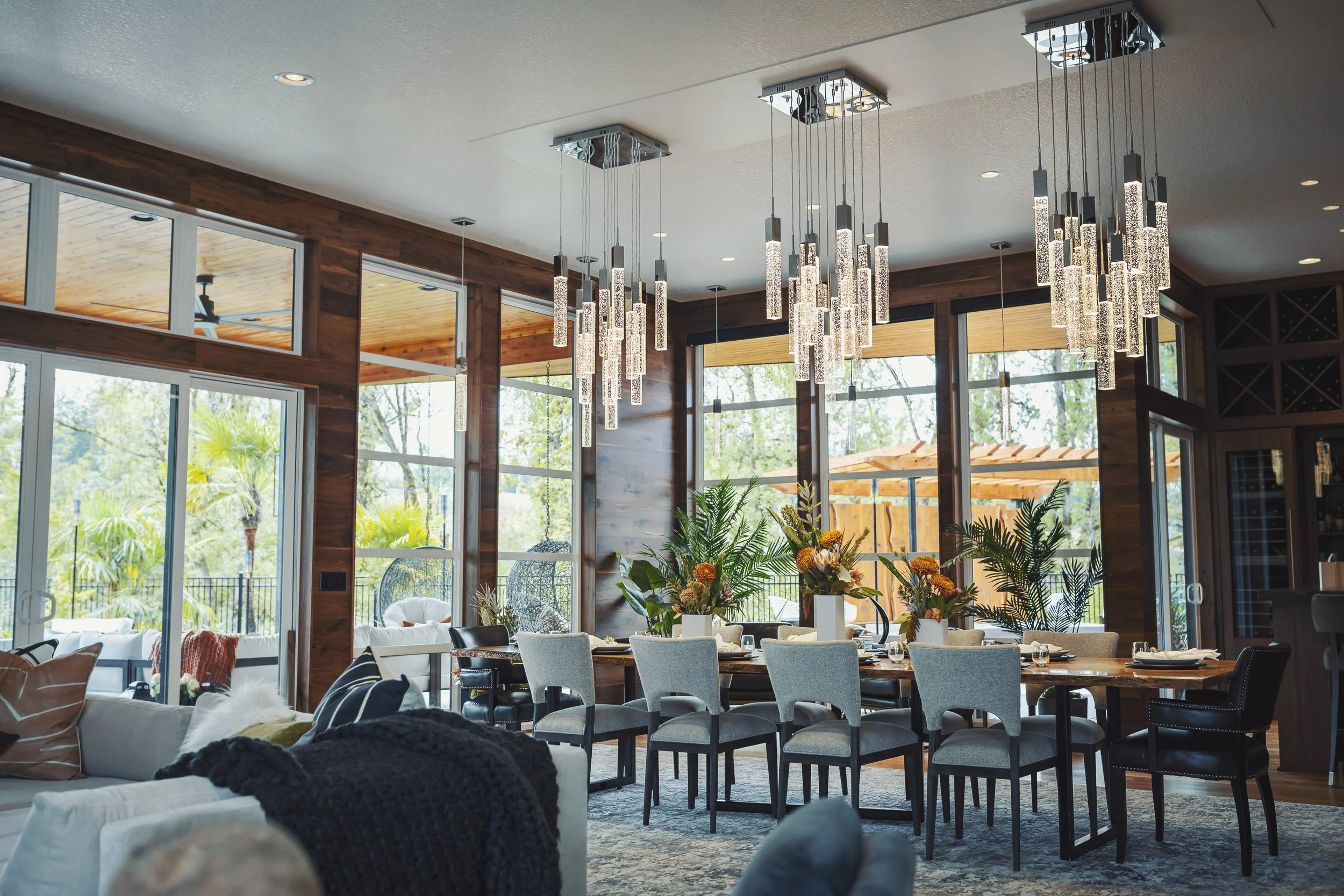 Interior view of a living and dining area with large windows, wooden accents, modern hanging chandeliers, a dining table with chairs, and decorative plants.