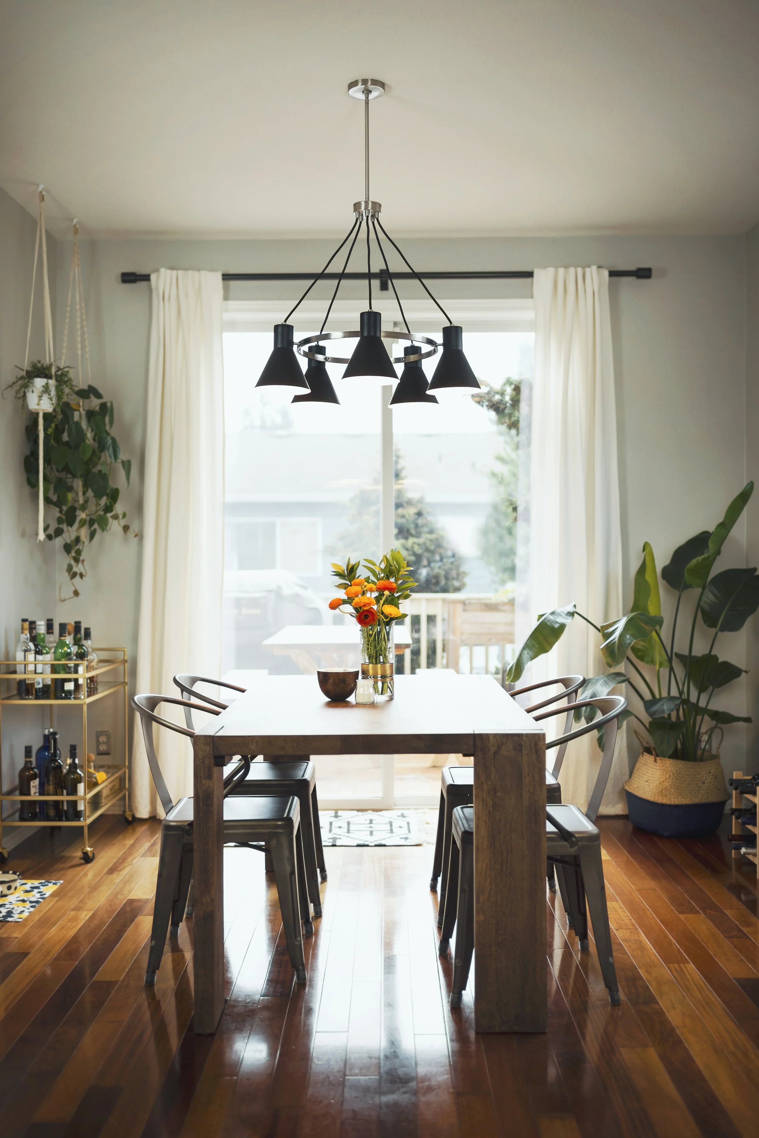 A dining room with a wooden table, six black chairs, a modern black chandelier, large window with white curtains, potted plants, and a wine rack.