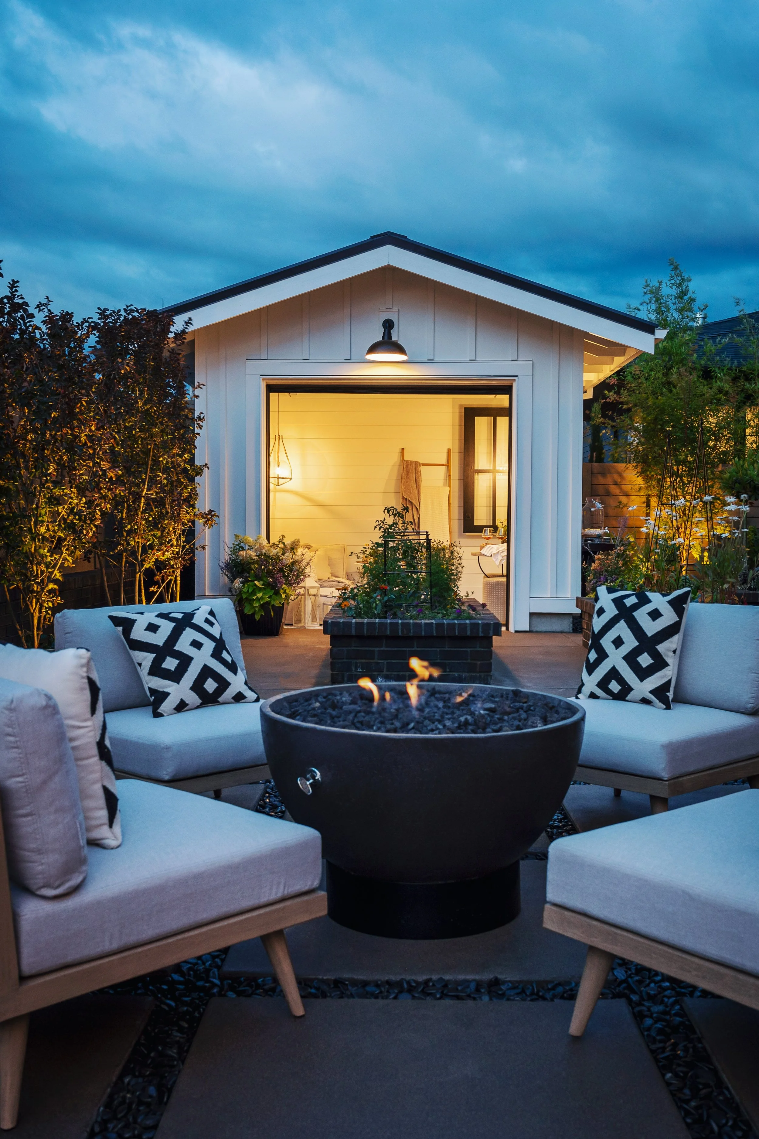 Outdoor patio at dusk with white cushioned seating, black-and-white patterned pillows, a fire pit, and a small house with a porch, warm interior lighting, and trees around.