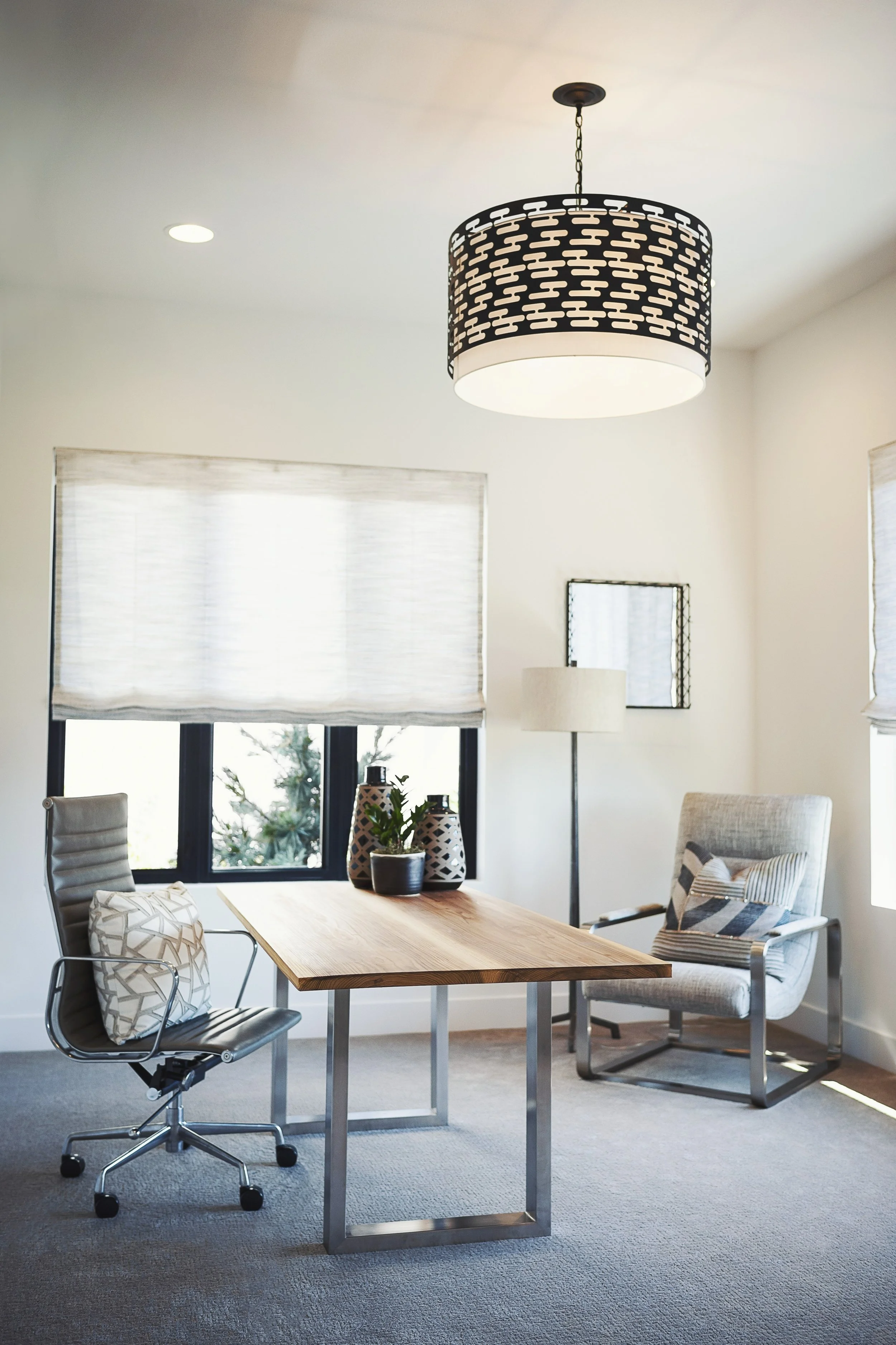 A modern home office with a wooden desk, office chair, and a lounge chair with a pillow. There is a window with a beige shades, a floor lamp, and wall art. A black and white pendant light hangs from the ceiling.